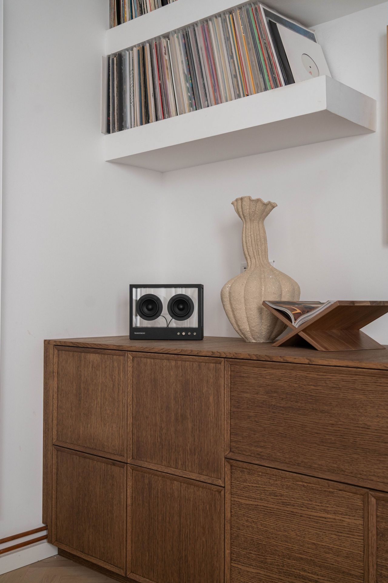 Kleiner transparenter Lautsprecher auf einem Holzschrank, daneben eine Vase und ein Schallplattenspieler mit Schallplatten im Hintergrund.