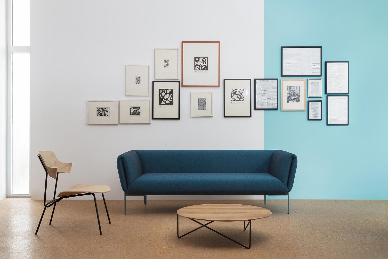 Blaues Impression Sofa von Prostoria mit Holzstuhl und rundem Couchtisch vor Bilderwand.