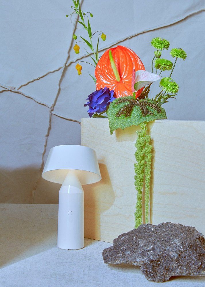 Weiße Bicoca Akkuleuchte / Tischleuchte von Marset neben Blumen und einem Stein.