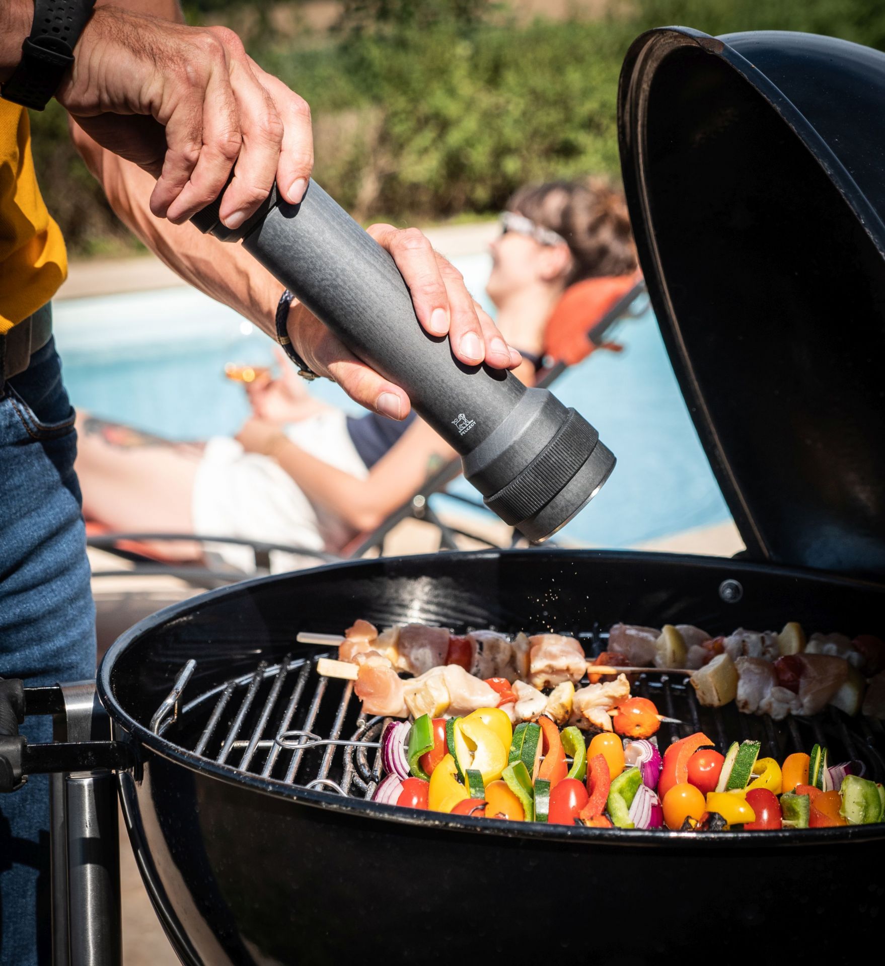 Mann würzt Grillspieße mit Peugeot BBQ Pfeffermühle über Holzkohlegrill im Garten.