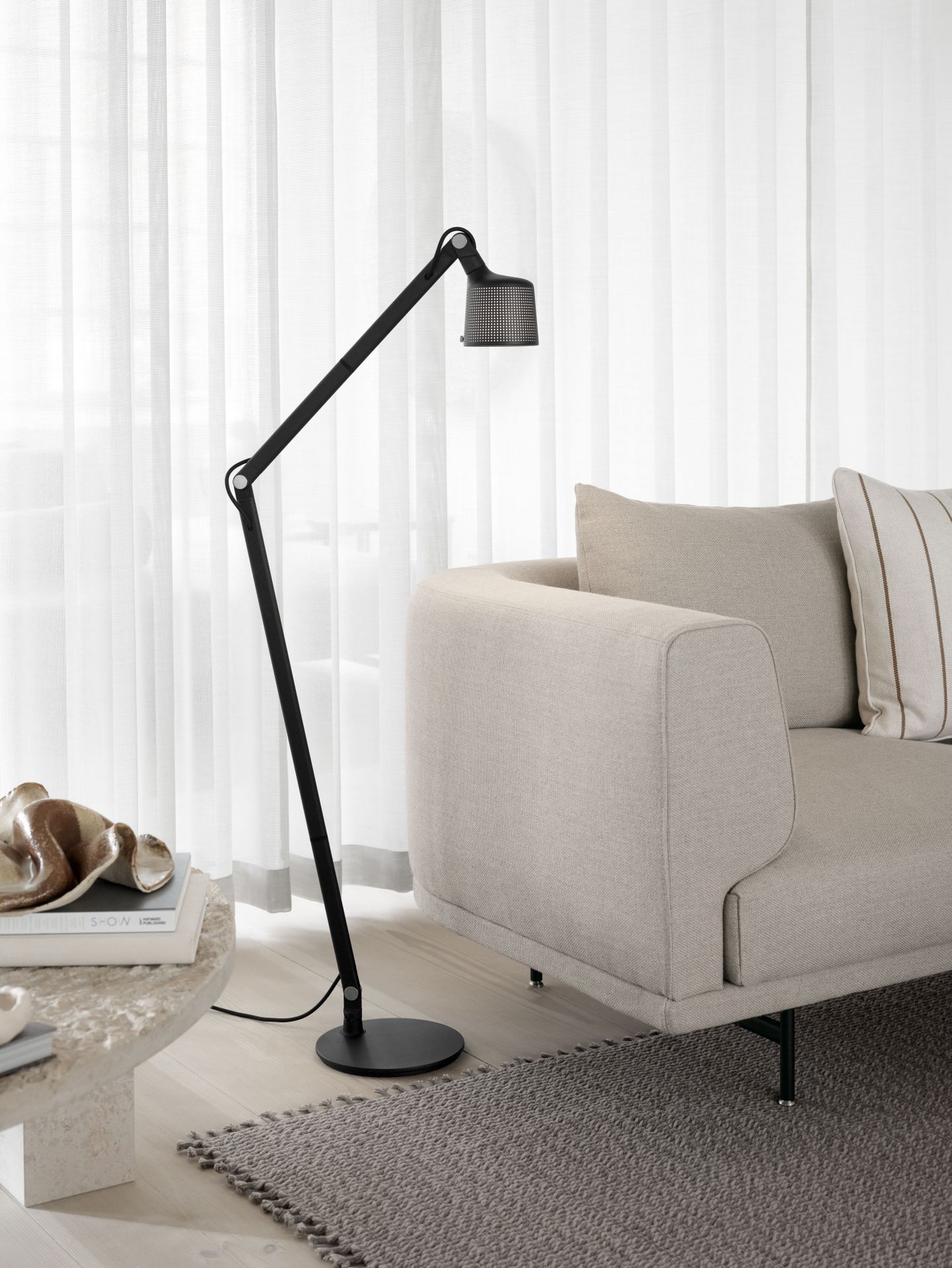 Schwarze Vipp Boden Leselampe neben beigem Sofa und Marmortisch mit Deko-Objekten vor hellem Vorhang.