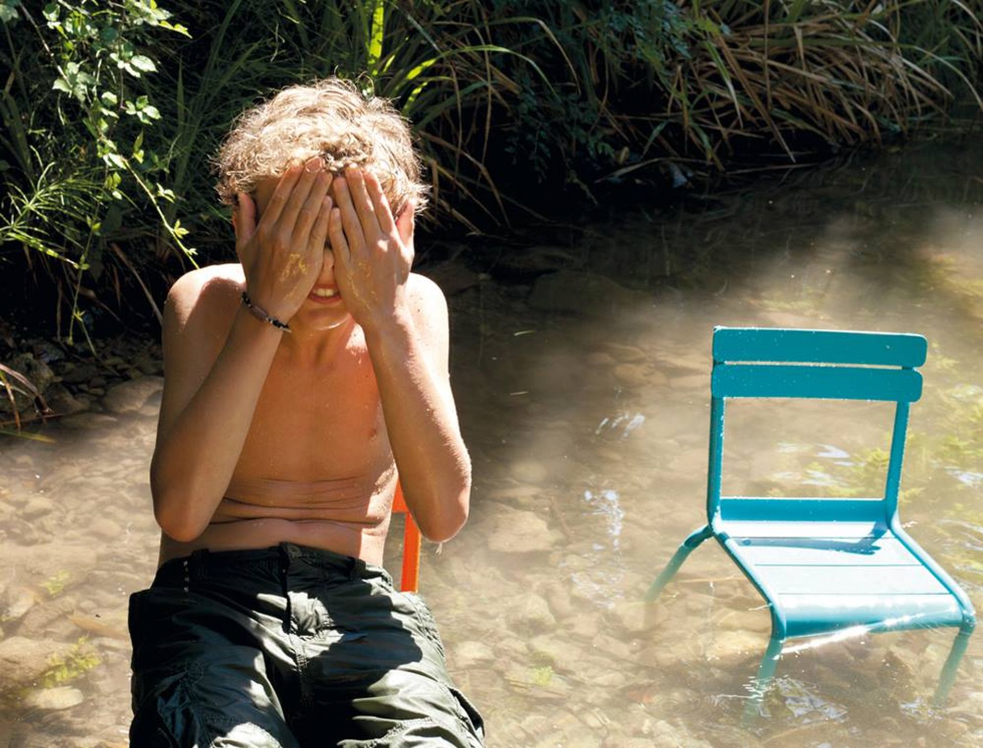 Junge sitzt im Wasser auf einem orangefarbenen Stuhl, daneben ein türkiser Luxembourg Kinderstuhl Outdoor von Fermob.
