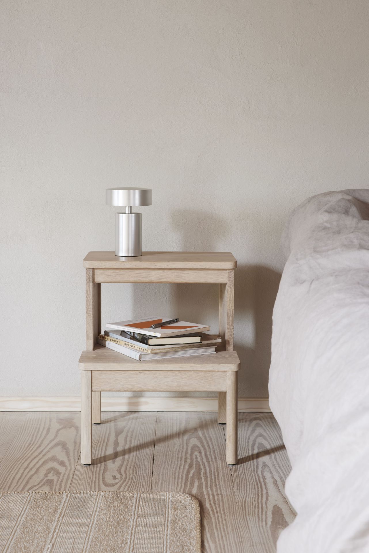 A Line Steighocker von Form & Refine aus hellem Holz als Nachttisch mit Lampe.