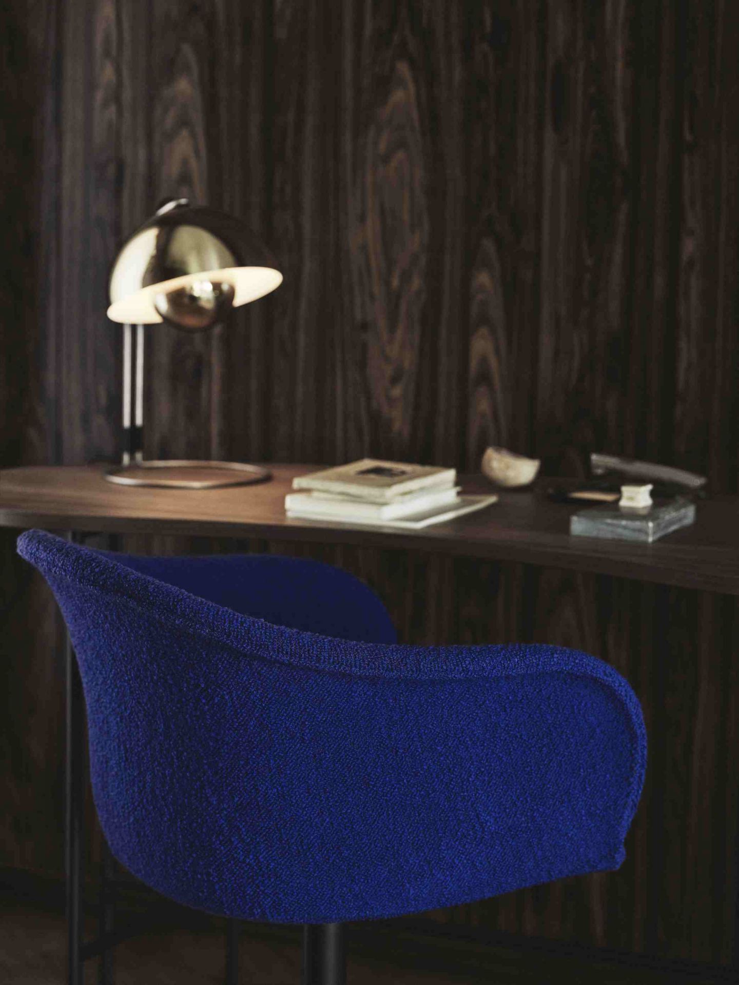 AndTradition Palette Schreibtisch mit Lampe und blauem Stuhl vor dunkler Holzvertäfelung, Bücher und Dekoration.