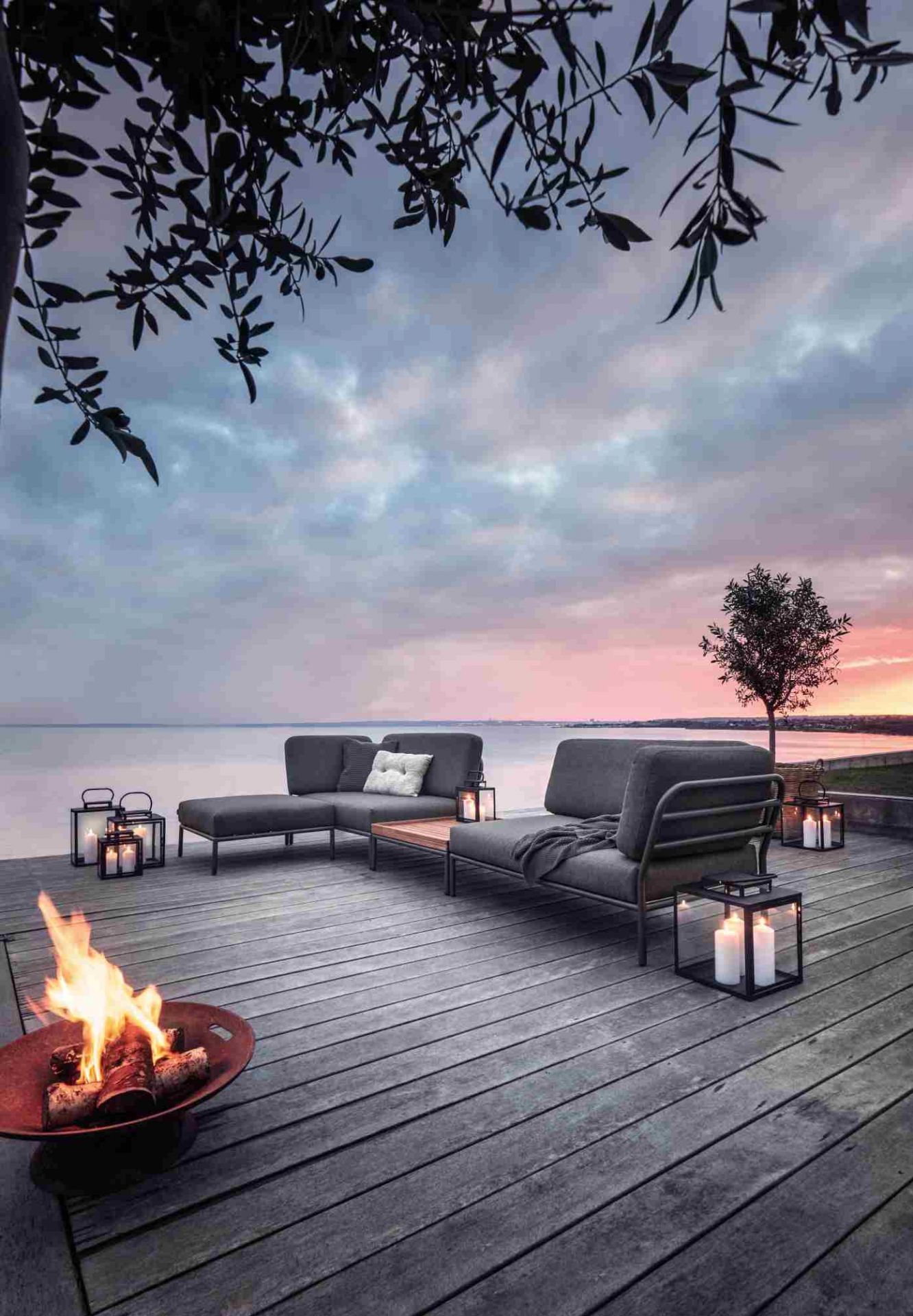 Houe Level Outdoor Sofa auf Holzterrasse mit Feuerstelle und Meerblick bei Sonnenuntergang.