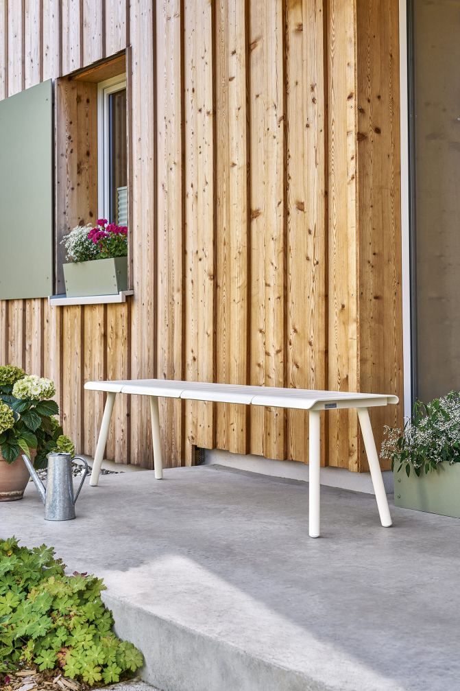 Weiße Vapio Bank von Weishäupl für den Außenbereich vor einer Holzwand mit Fenster und Blumenkästen.