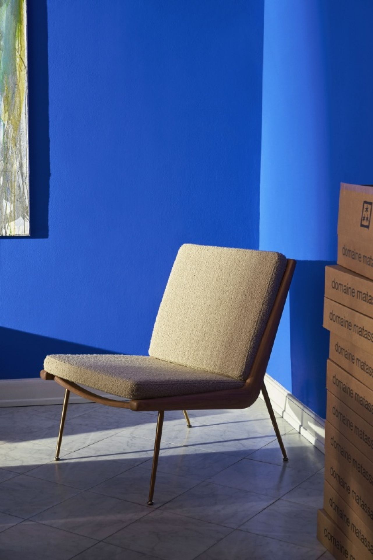 AndTradition Boomerang Lounge Chair HM1 Sessel mit beigem Stoffbezug und filigranem Metallgestell vor blauer Wand.