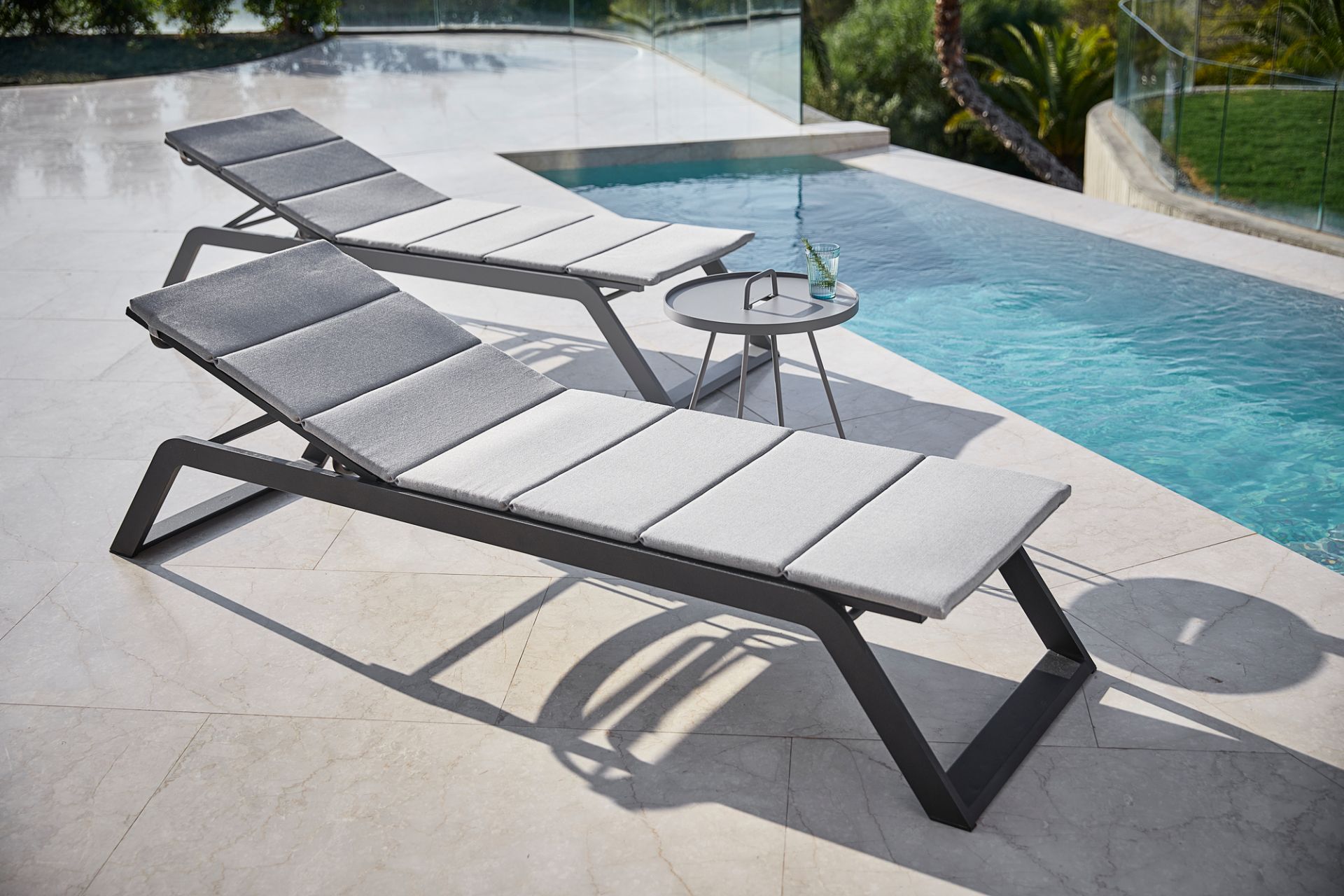 Zwei graue Cane-Line Sonnenliegen mit Polsterauflagen am Pool, moderne Gartenmöbel für den Außenbereich.