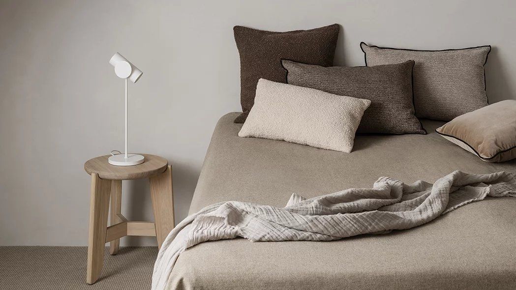 Eli Stool Sitzhocker von Blomus aus Holz neben einem Bett mit Kissen und Lampe.