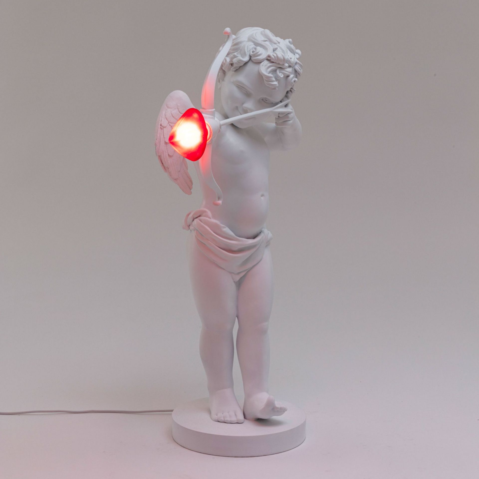 Cupid Lampe von Seletti: Tischleuchte in Form eines weißen Amor mit rotem Herz-Pfeil.