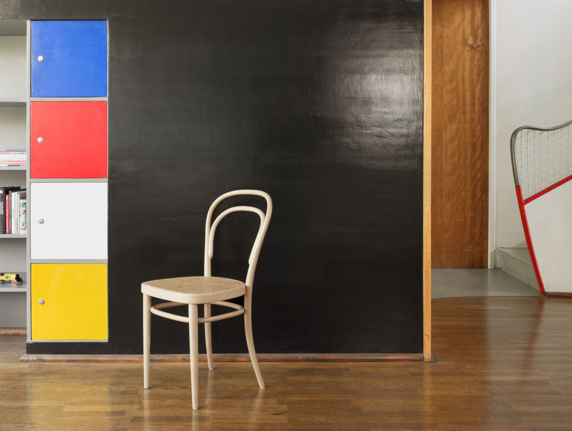 214 Bugholzstuhl, klassischer Wiener Kaffeehausstuhl mit Rohrgeflecht Sitzfläche vor schwarzer Wand und bunten Schränken.