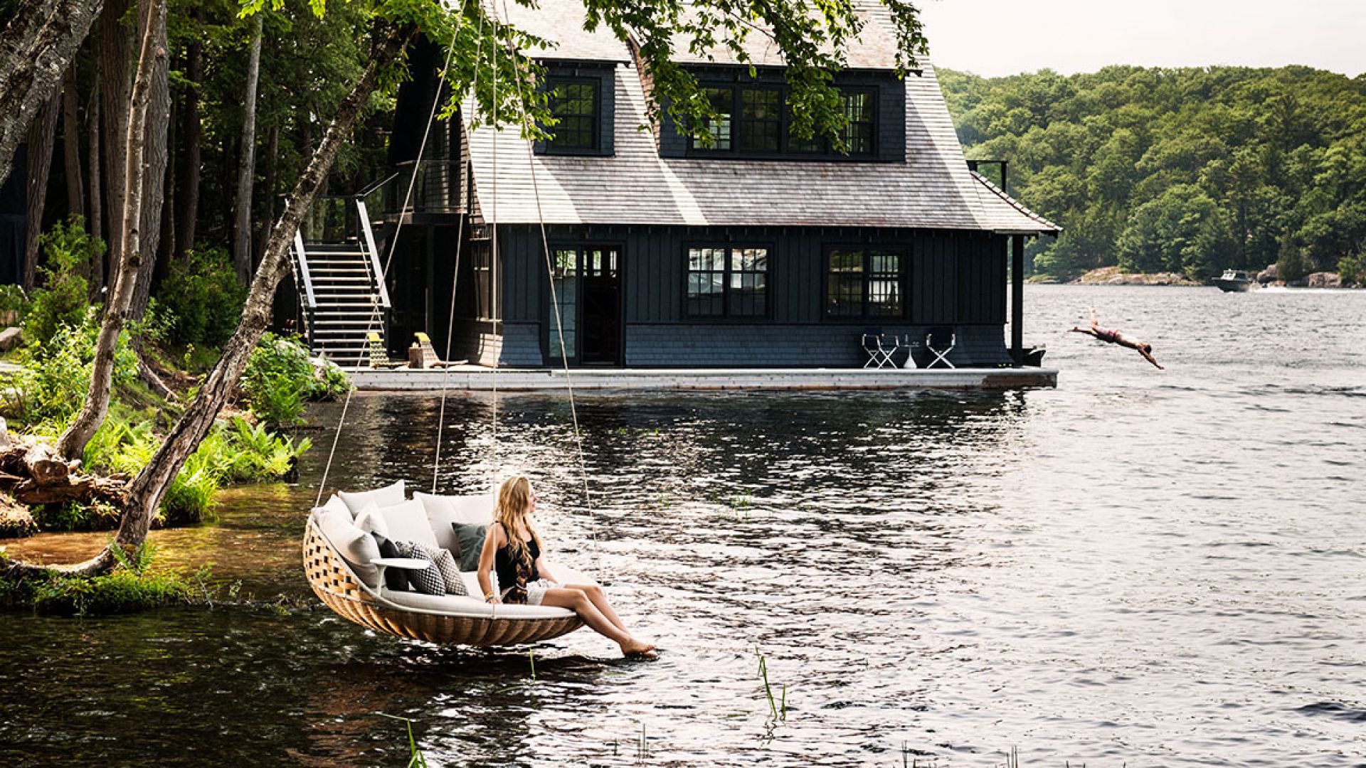Frau entspannt auf der Dedon Swingrest Hängelounge mit Kissen am See, während eine Person ins Wasser springt.