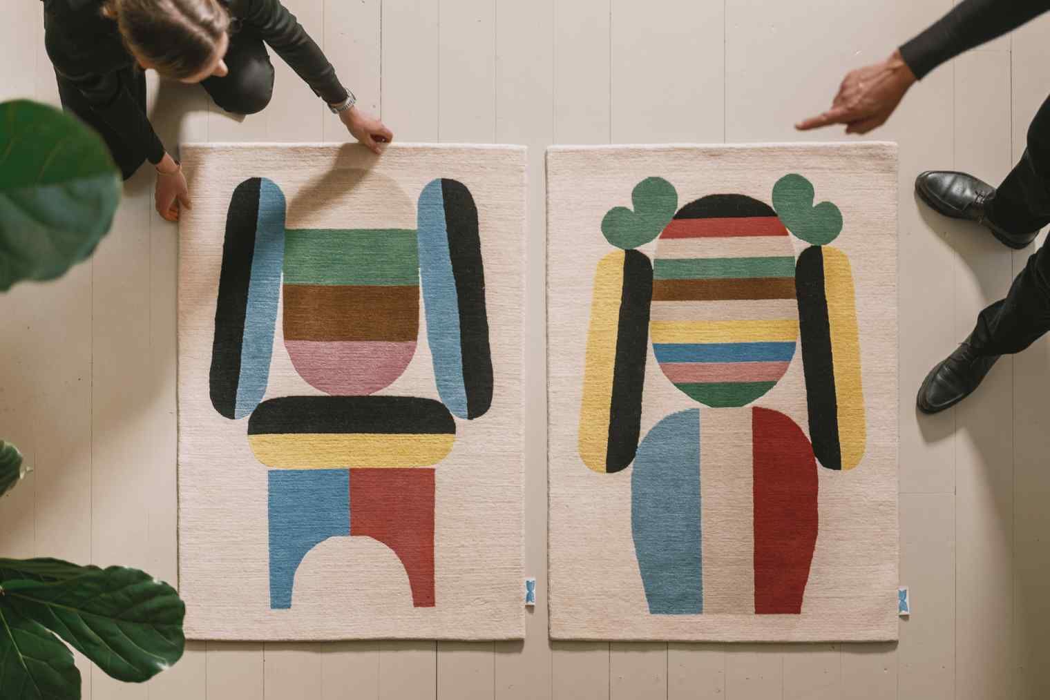 Zwei Bela Teppiche von Designercarpets mit abstraktem, farbenfrohem Muster auf hellem Untergrund, präsentiert von zwei Personen.