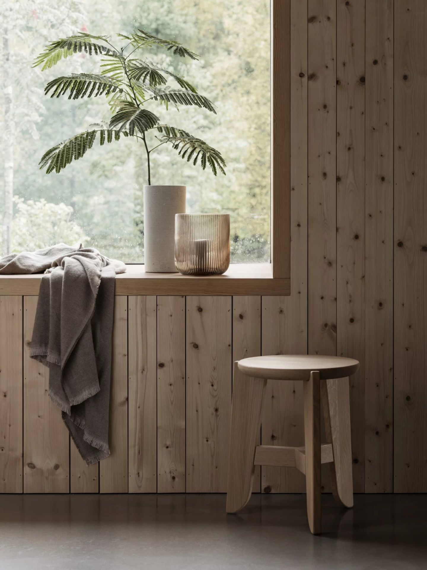 Eli Stool Sitzhocker von Blomus aus hellem Holz vor einer Holzwand und Fenster.