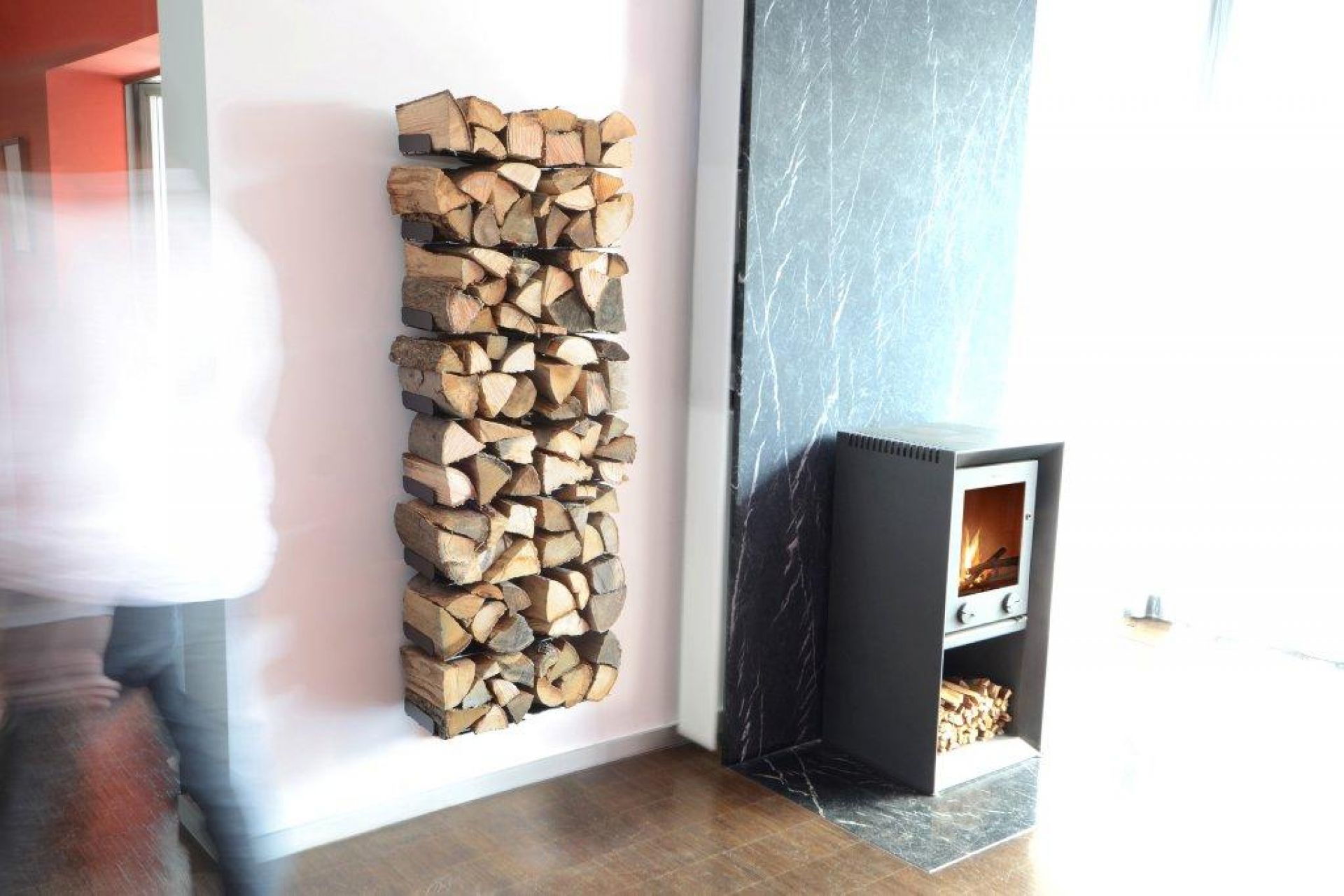 Wooden Tree Kaminholzregal Radius, wandmontiert und gefüllt mit Brennholz, neben einem Kaminofen im modernen Wohnzimmer.