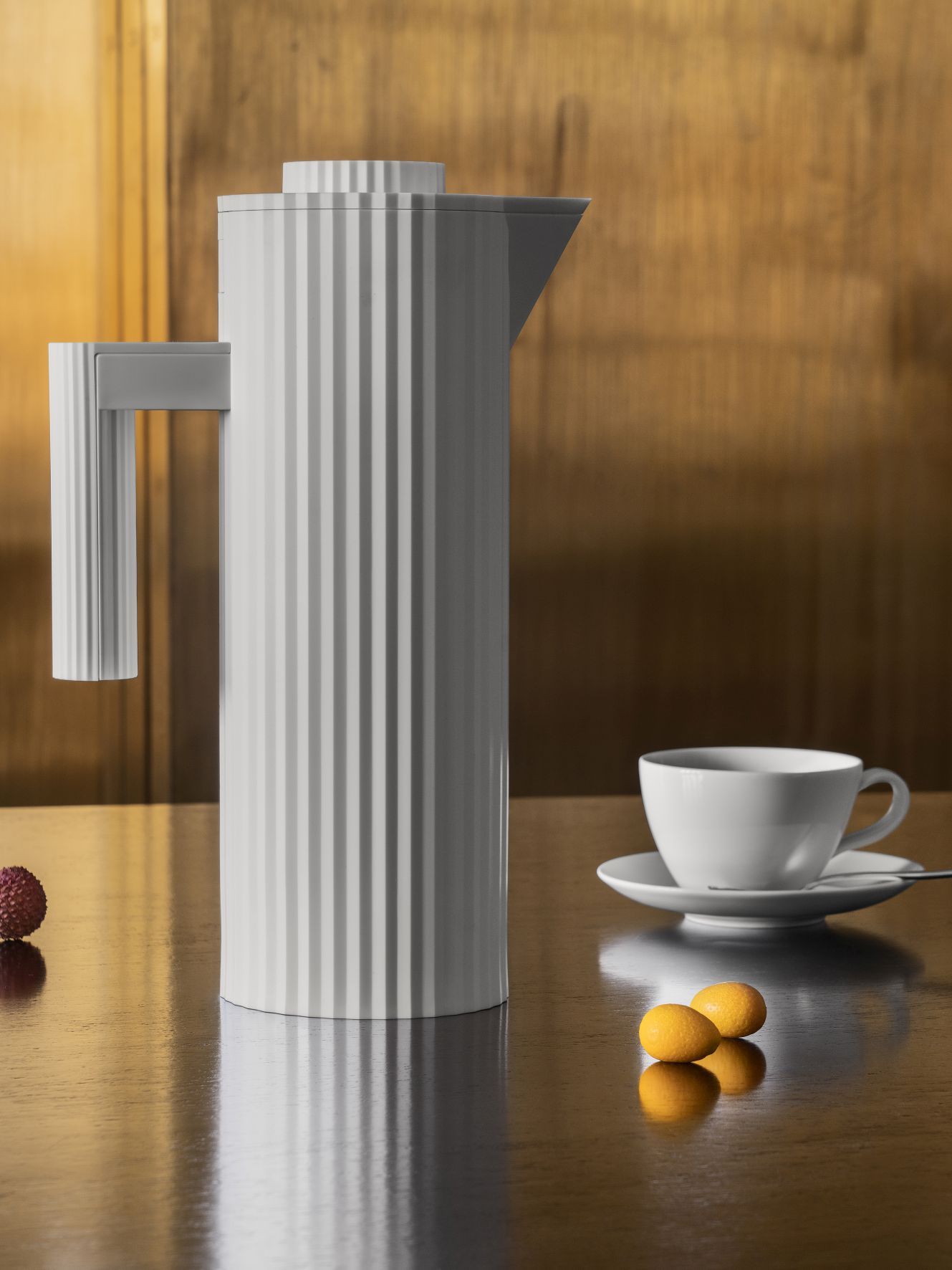 Graue Alessi Isolierkanne MDL12 mit Plissee-Design neben Kaffeetasse und Früchten auf glänzender Oberfläche.