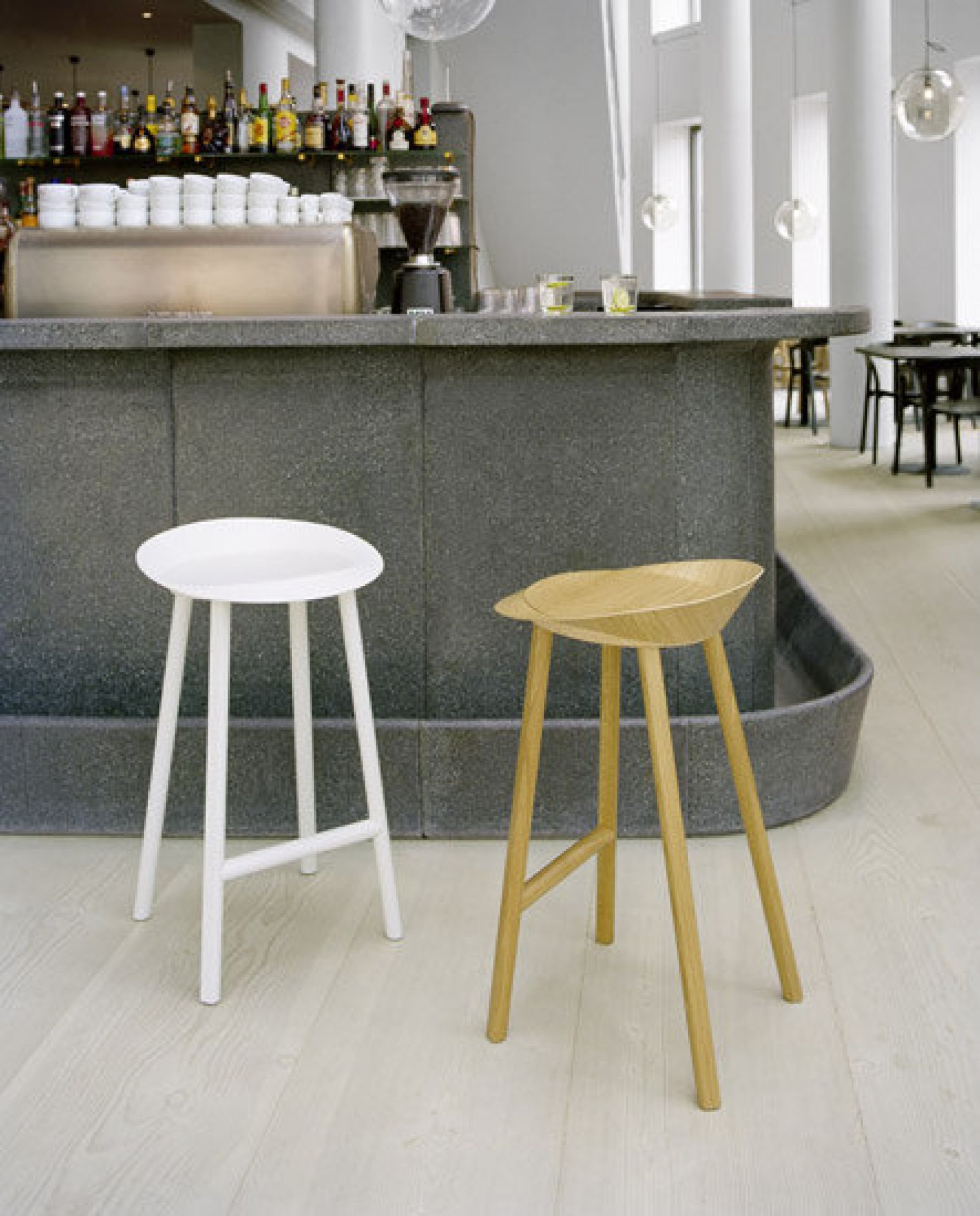 Zwei moderne Barhocker aus Holz, einer weiß lackiert, der andere naturfarben, vor einer Bar.