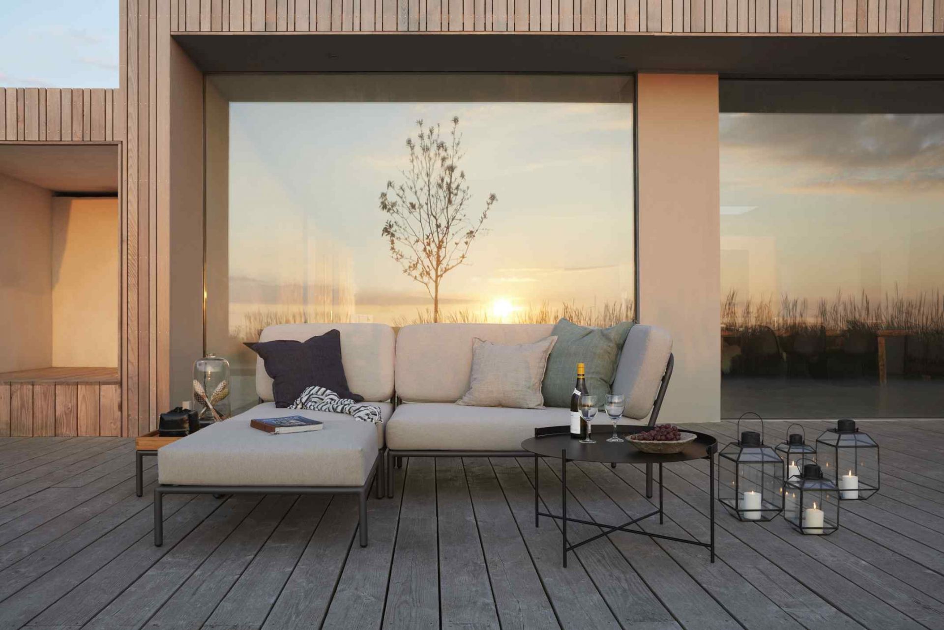 Level Sofa Outdoor von Houe auf Holzterrasse mit Tisch, Windlichtern und Blick auf den Sonnenuntergang.