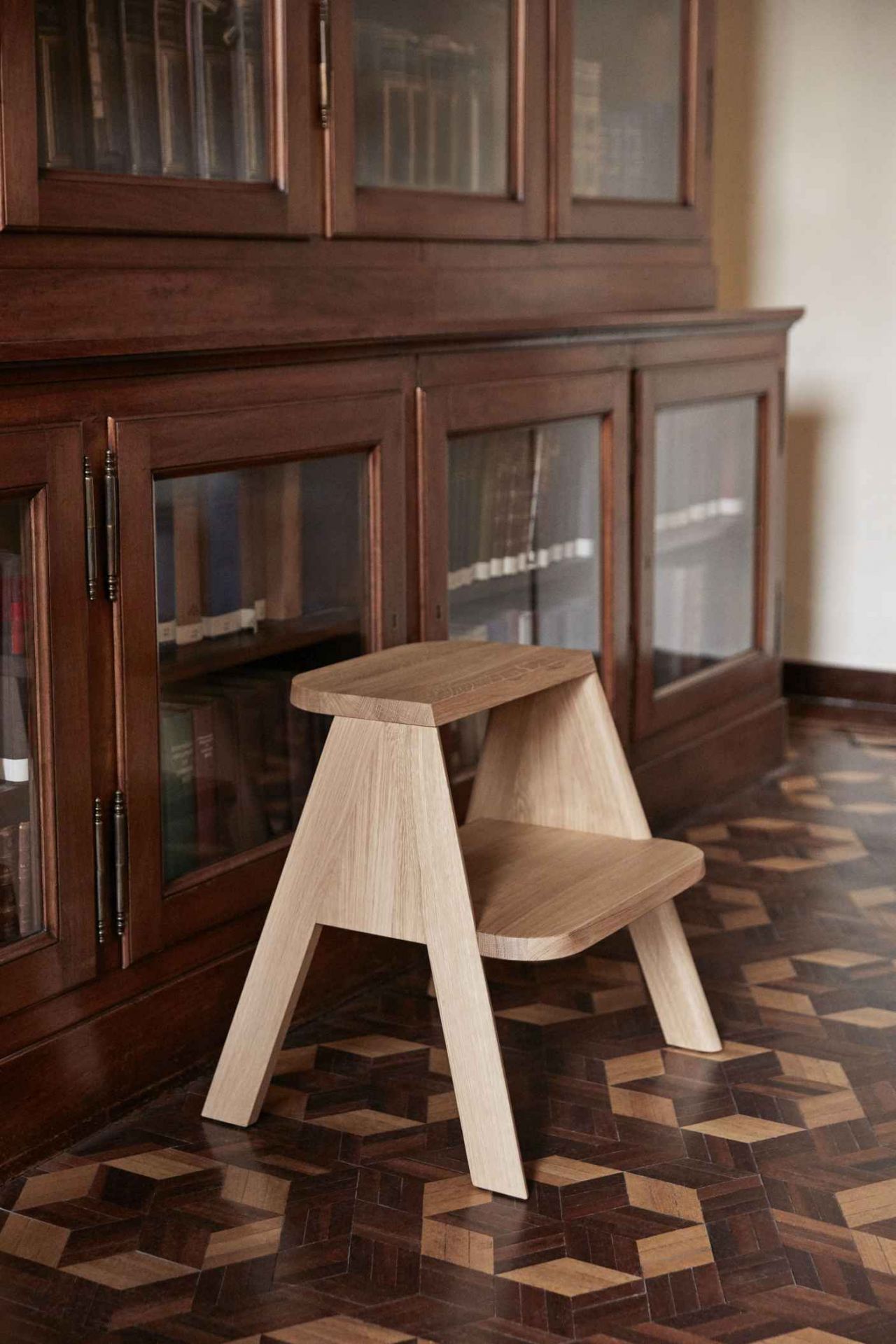 Butler Hocker von Hay aus hellem Eichenholz vor einem dunklen Holzschrank und Parkettboden.