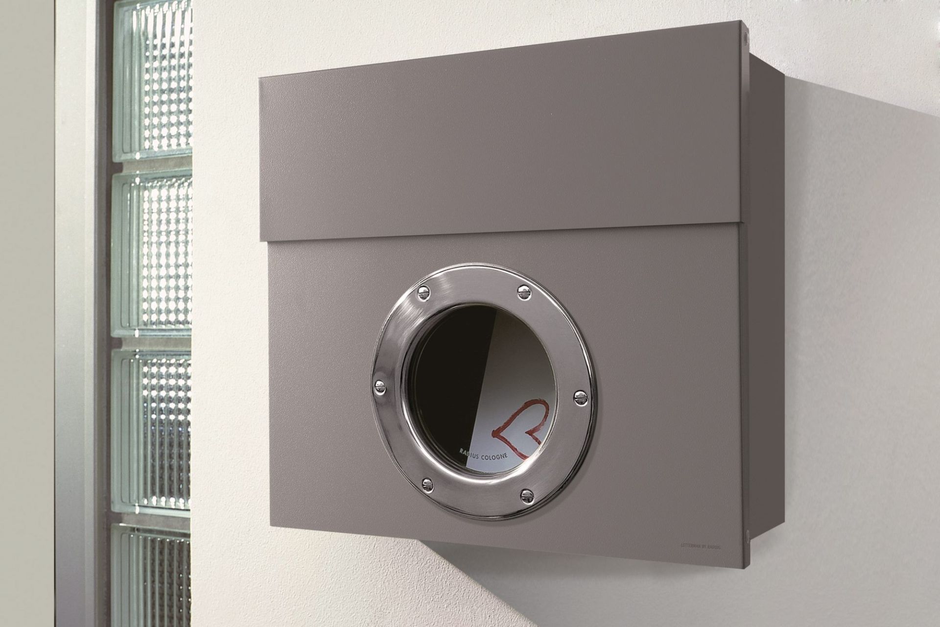Grauer Letterman Briefkasten Radius I mit Sichtfenster aus Edelstahl an einer weißen Wand.