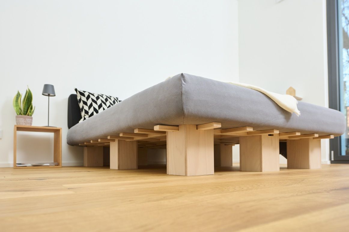 Tojo Cube Nachttisch aus Holz neben einem minimalistischen Bett mit grauer Matratze und gemustertem Kissen.