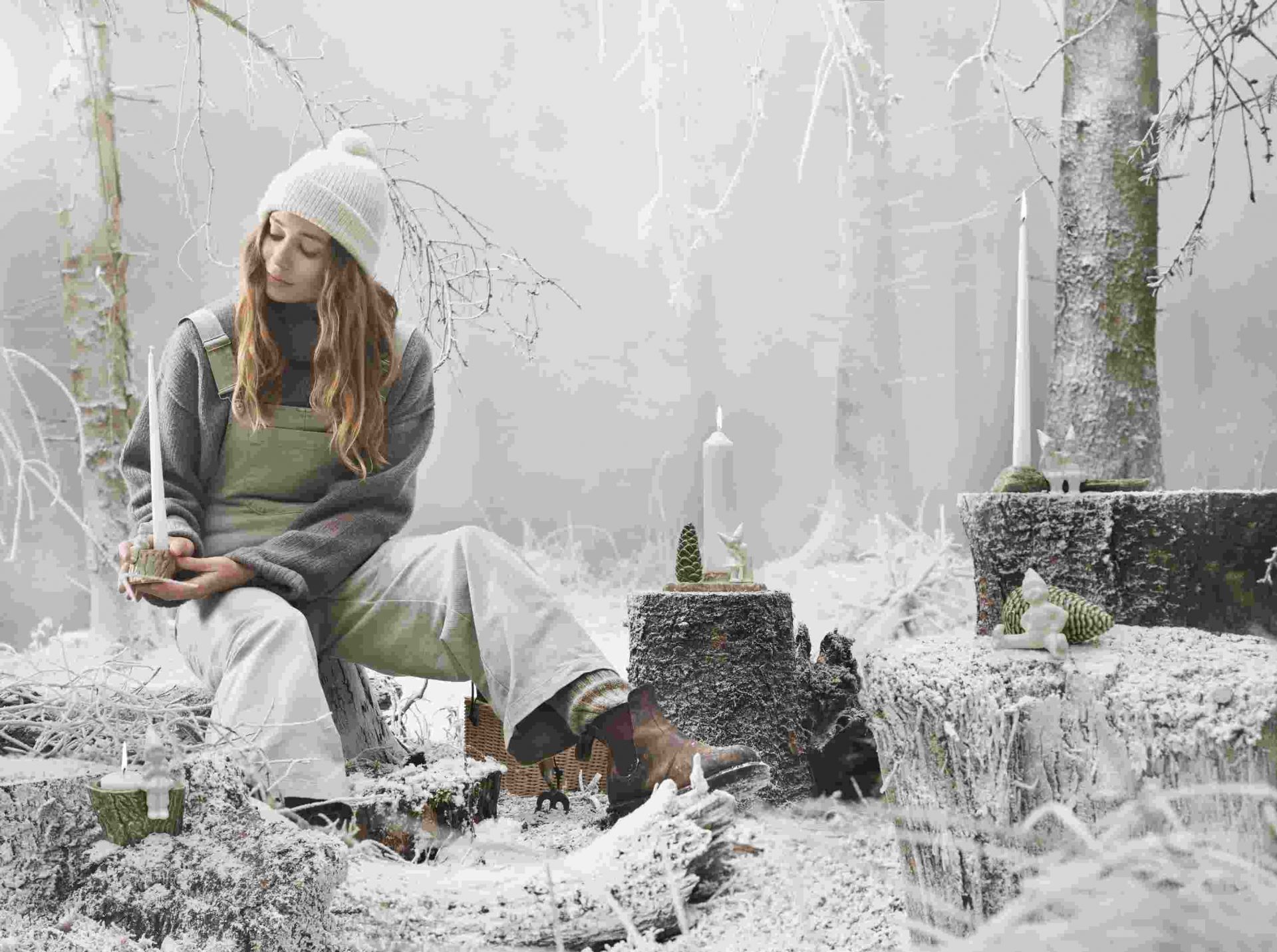 Frau im Winterwald mit Kähler Christmas Tales Julekram Kerzenständer und weiterer Weihnachtsdekoration auf Baumstümpfen.