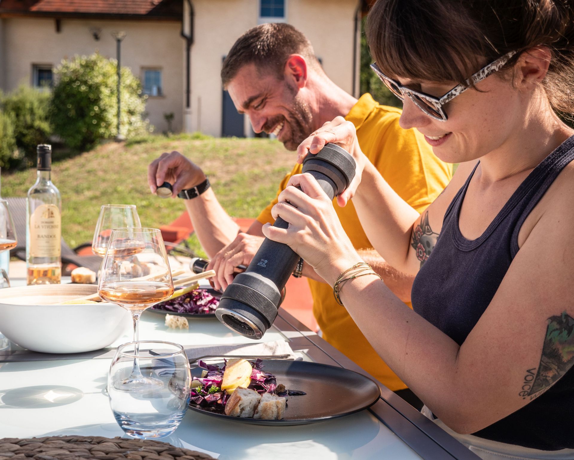 Draufsicht: Frau würzt Salat mit Peugeot BBQ Pfeffermühle graphit, Outdoor-Szene mit Wein.