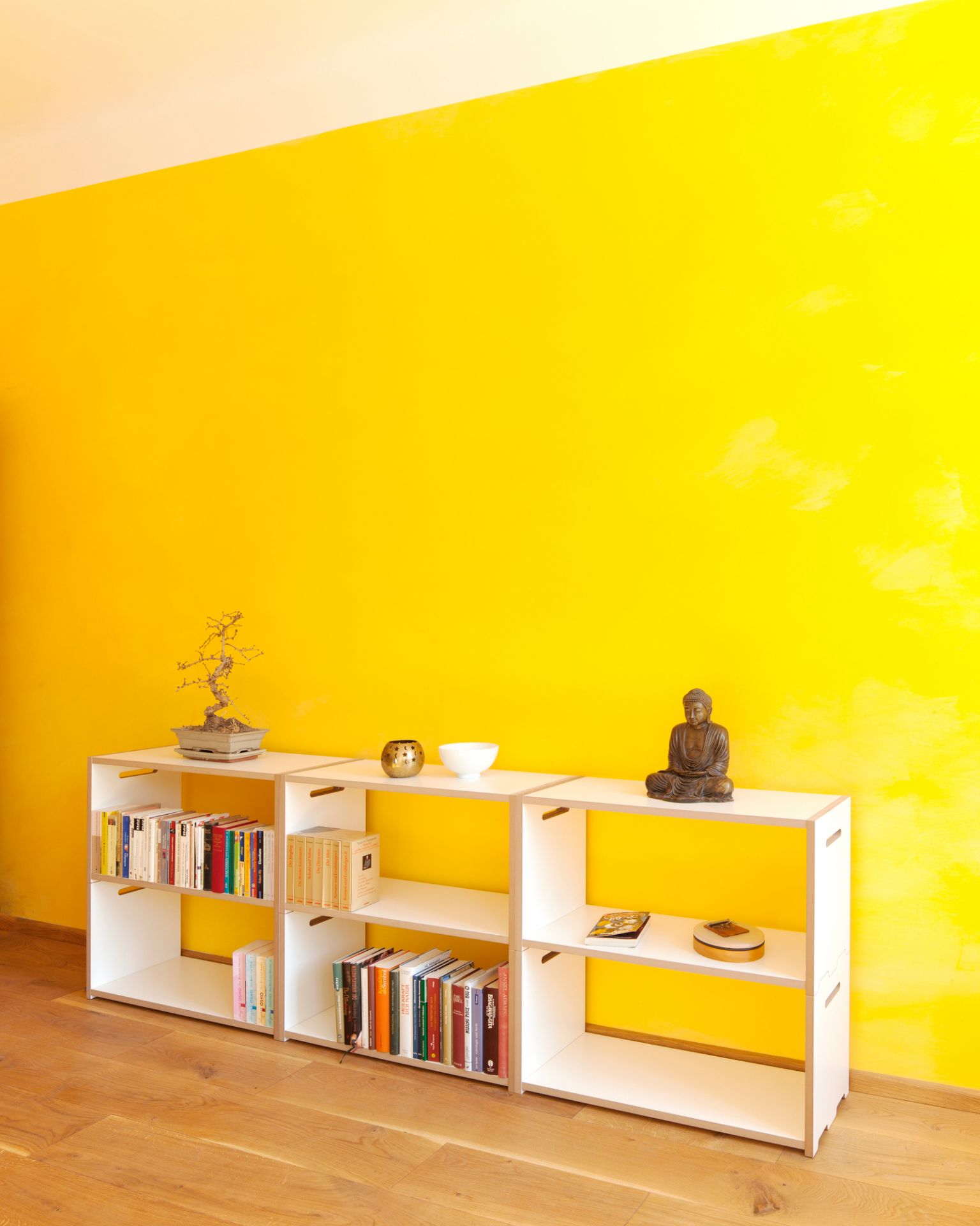 Weißes Tojo Hochstapler Regal Anbaumodul vor gelber Wand, dekoriert mit Büchern, Bonsai und Buddha-Statue.