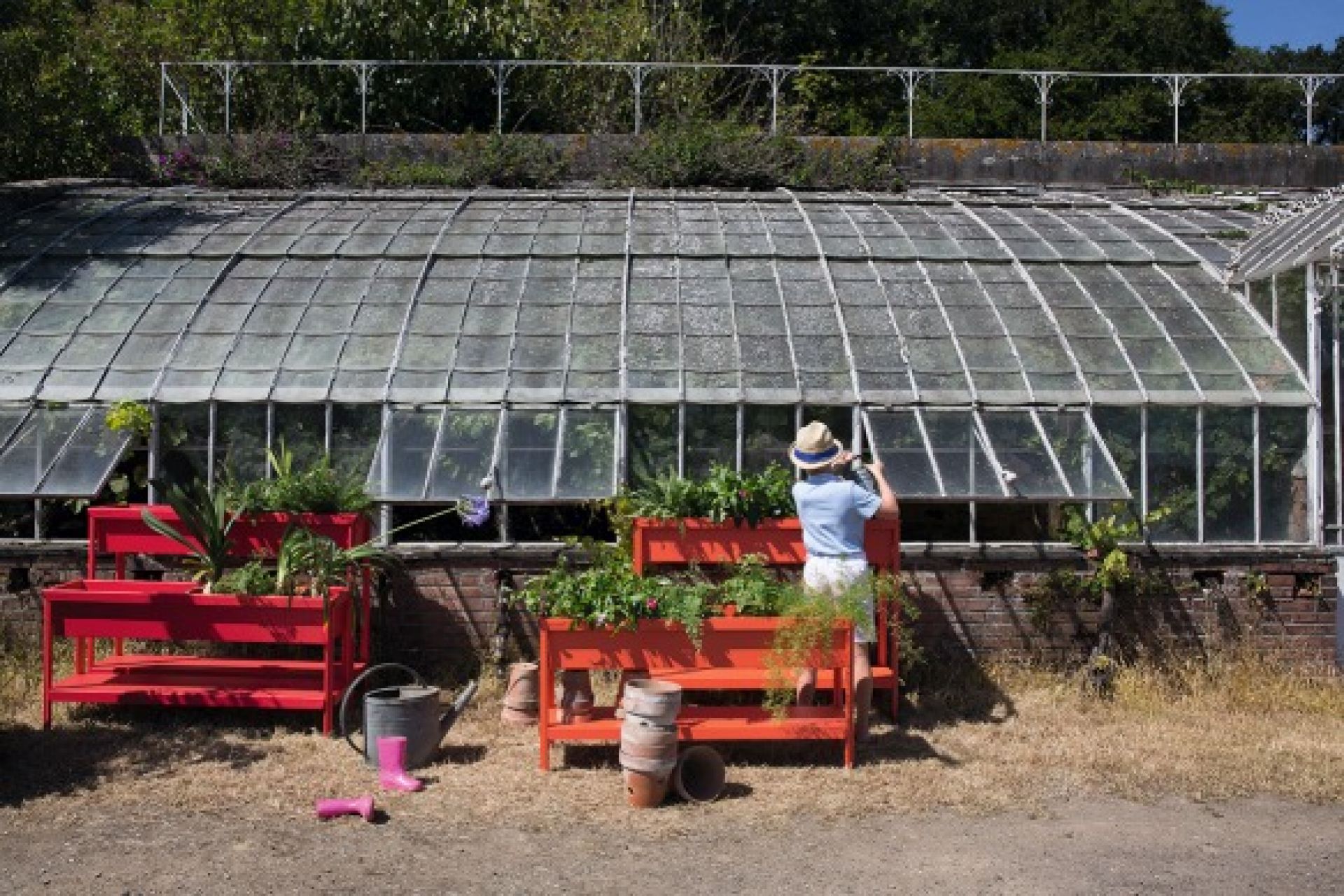 Roter Fermob Terrazza Blumenkasten vor Gewächshaus mit Person, die gießt, für Garten und Terrasse.