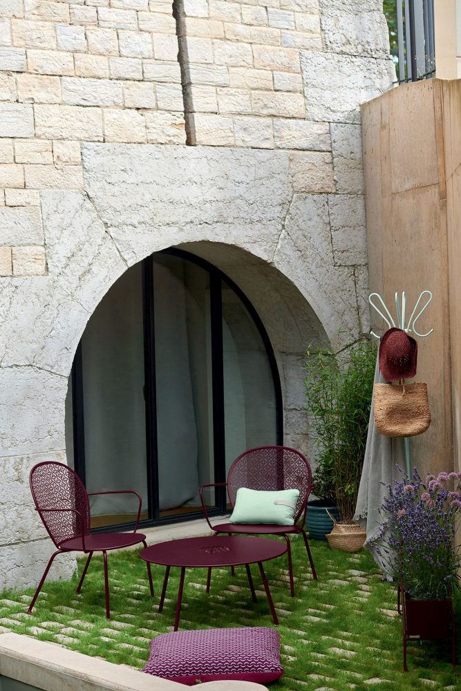 Fermob Lorette Sessel und Tisch in Pflaume auf Rasenfläche vor Steinmauer. Outdoor-Möbel für Garten und Terrasse.