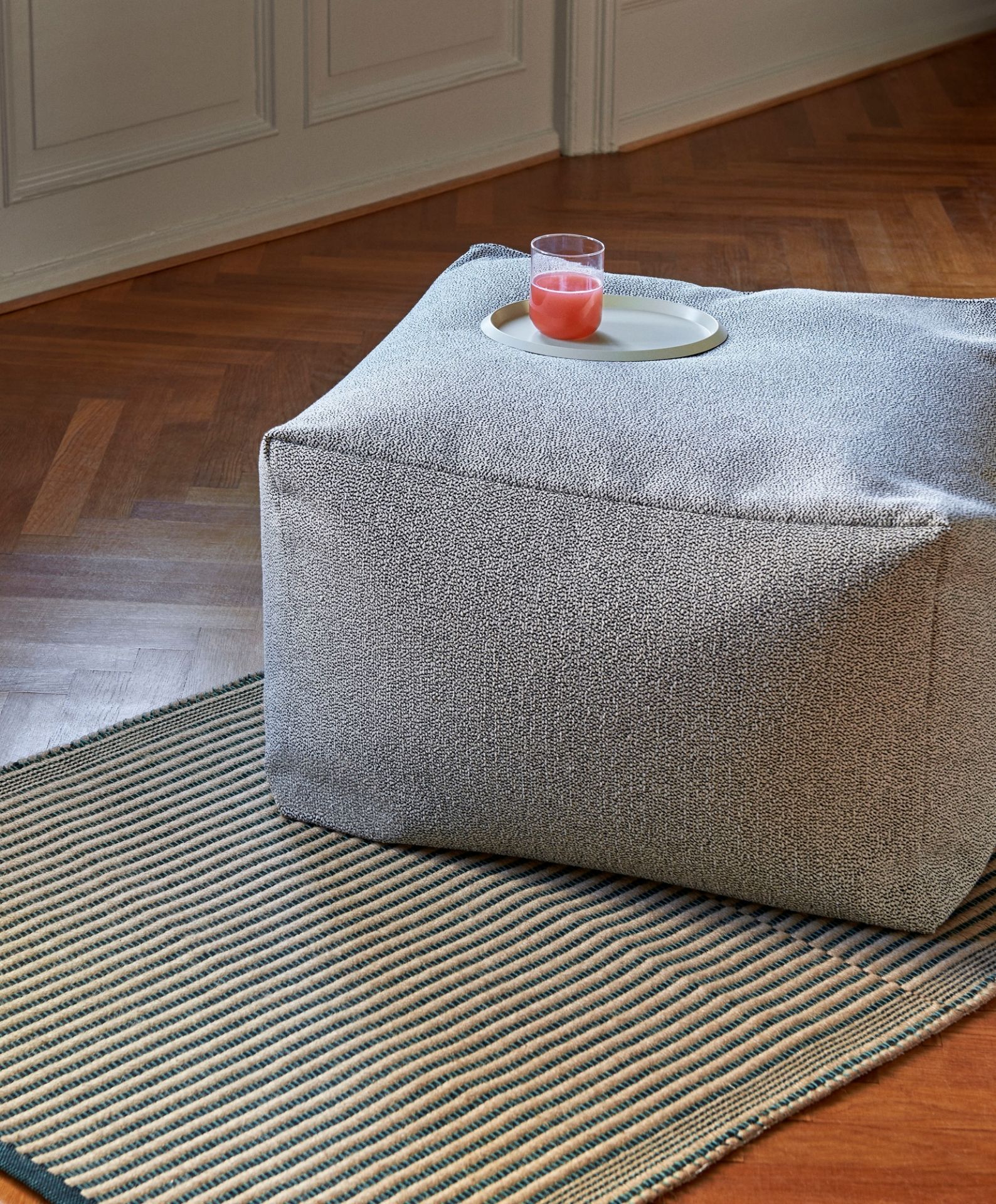 Nahaufnahme des Hay Tapis Teppichs mit Streifenmuster und einem grauen Sitzhocker mit Getränk.