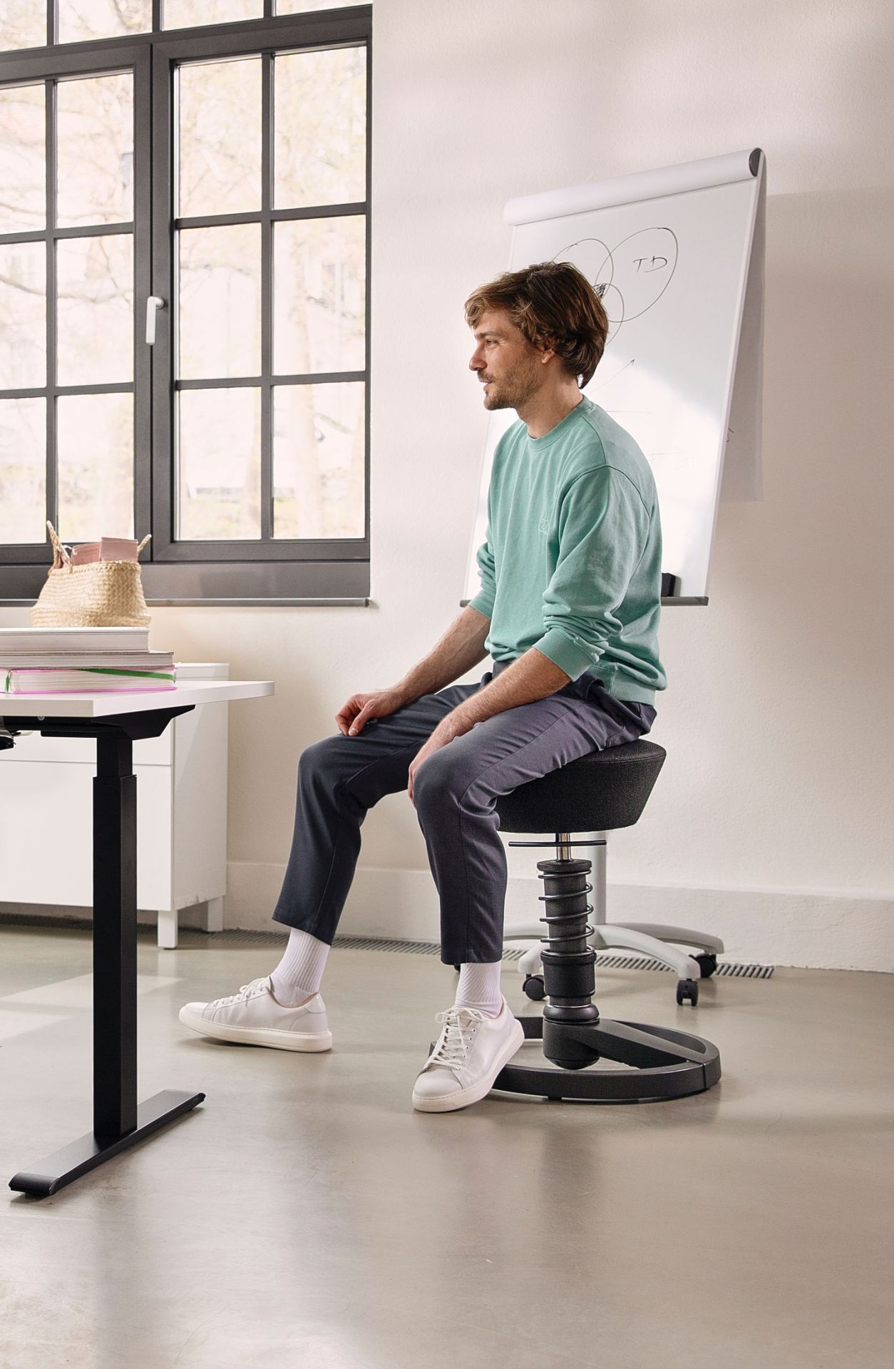 Mann sitzt auf schwarzem, höhenverstellbarem Bürohocker mit Spiralfeder vor Flipchart und Schreibtisch.
