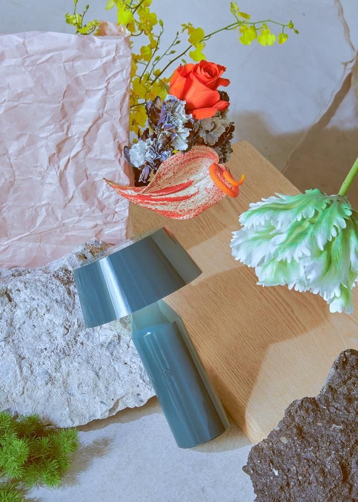 Blaue Bicoca Akkuleuchte von Marset als Tischleuchte, umgeben von Blumen und Steinen.