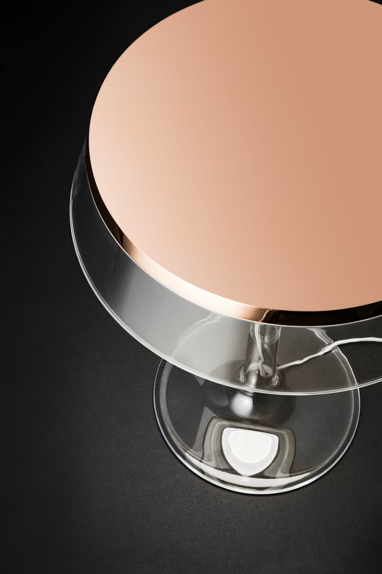 Detailaufnahme der Bon Jour Tischleuchte von Flos mit roségoldenem Schirm und transparentem Fuß auf schwarzem Hintergrund.