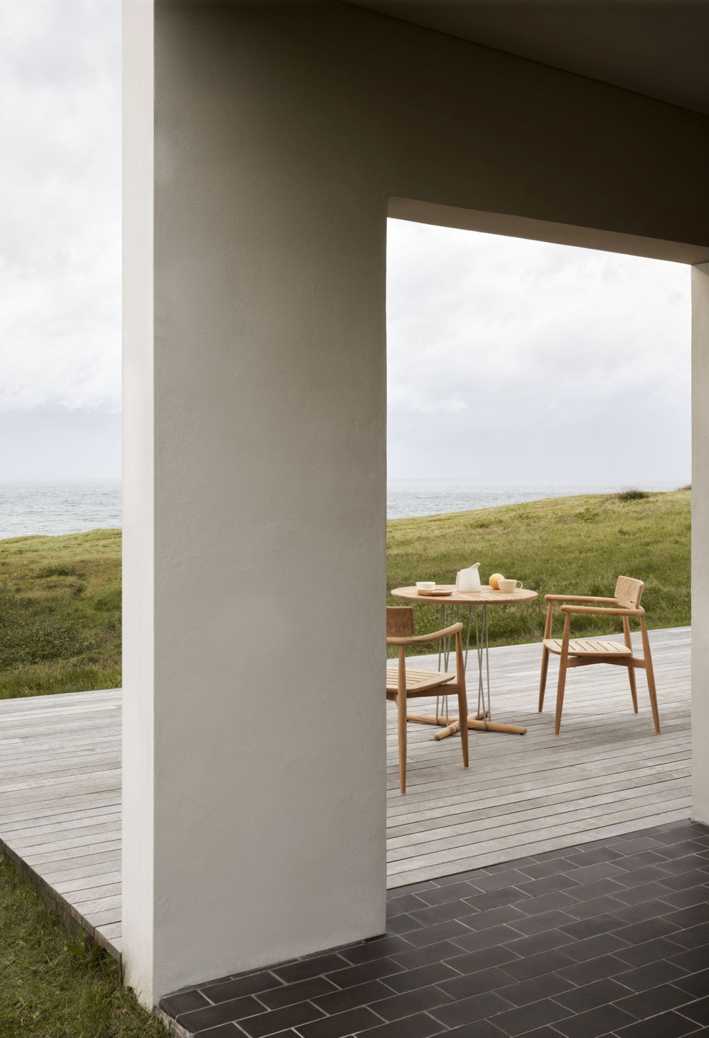 E008 Embrace Outdoor Stühle von Carl Hansen & Søn mit Tisch auf Holzterrasse am Meer.