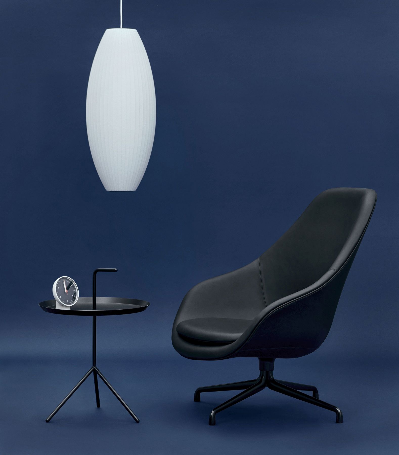 Hay Tischuhr auf Beistelltisch neben schwarzem Sessel und Lampe vor blauem Hintergrund.