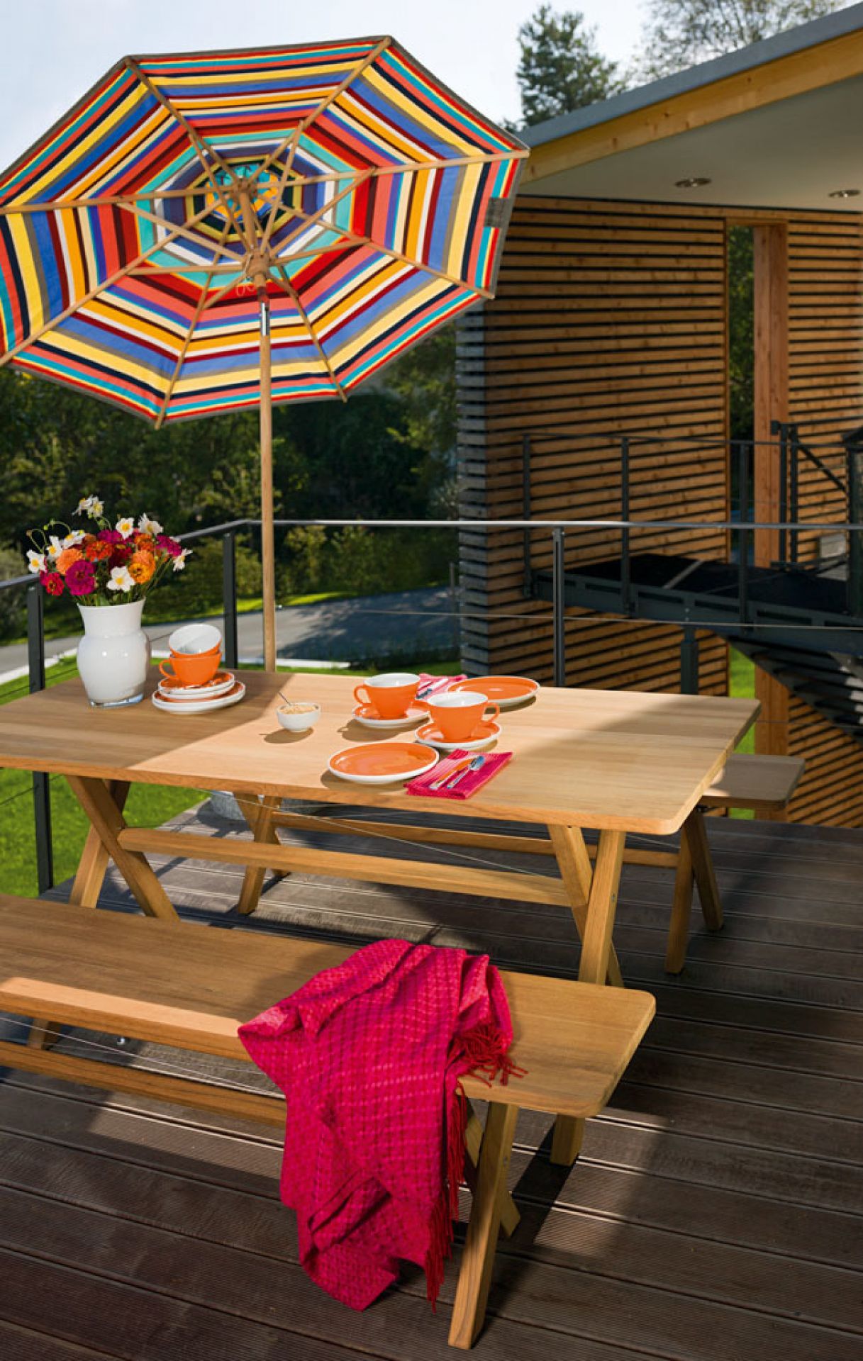 Weishäupl Cross Bank klappbar auf Terrasse mit Tisch, Bänken, Geschirr und buntem Sonnenschirm.