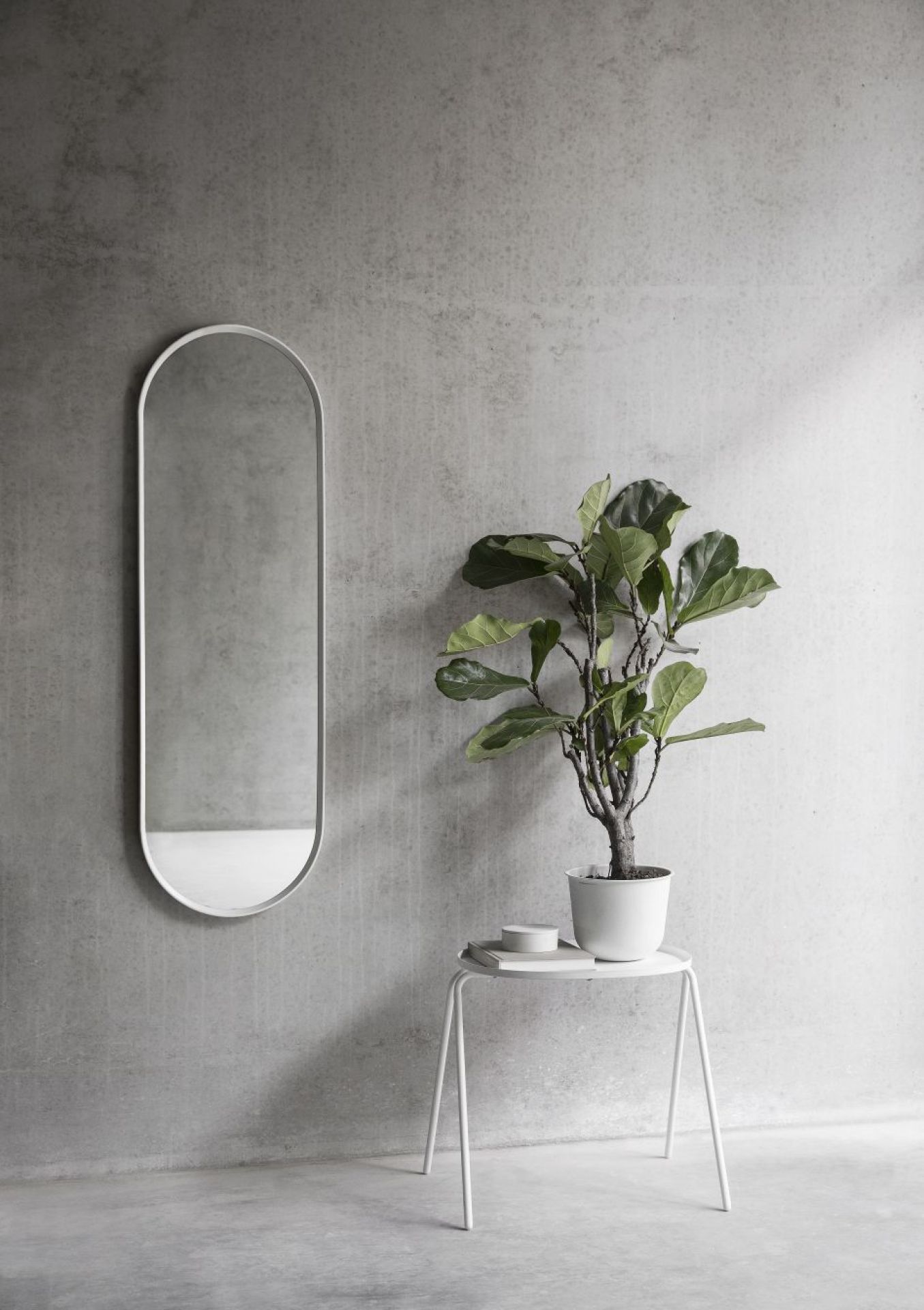 Norm Wandspiegel Oval von Audo mit weißem Rahmen an grauer Wand, neben Pflanze.