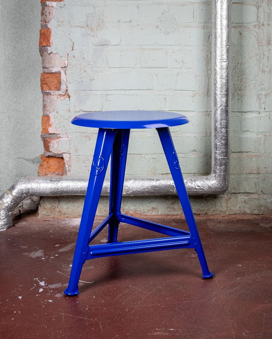 Blauer Rowac Schemel IS 48 cm Hocker Blueprint Edition vor einer Backsteinwand und Rohrleitung.