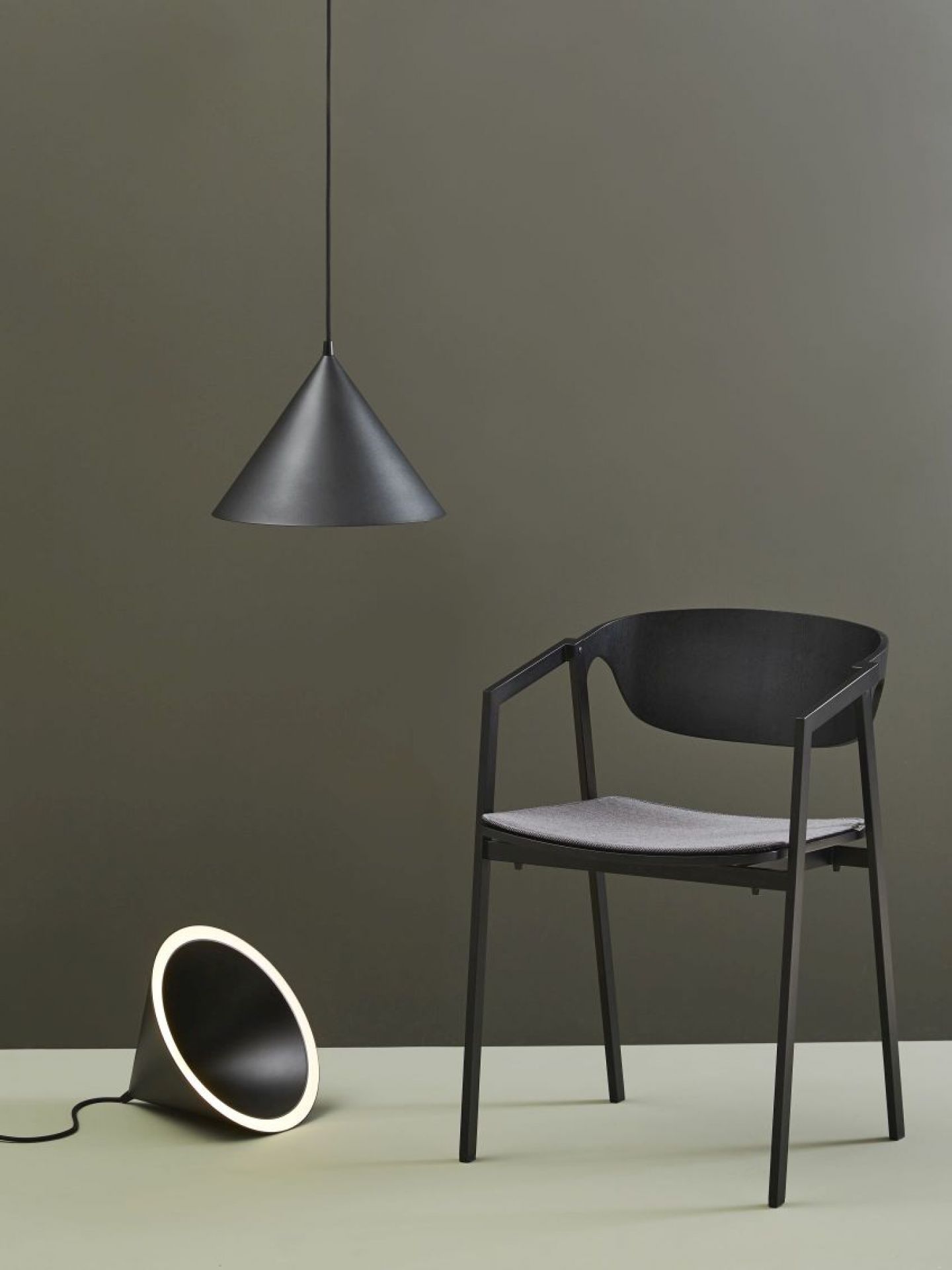 Annular Pendant Pendelleuchte von Woud, schwarz, kegelförmig, neben einer Tischleuchte und einem Stuhl.