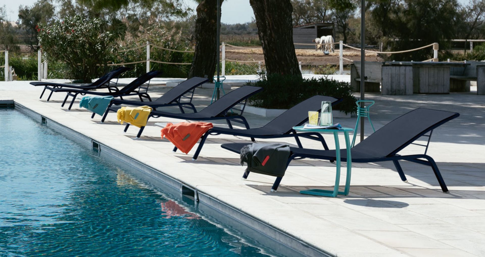 Mehrere blaue Alizé Sonnenliegen von Fermob stehen an einem Pool, mit farbigen Handtüchern dekoriert.