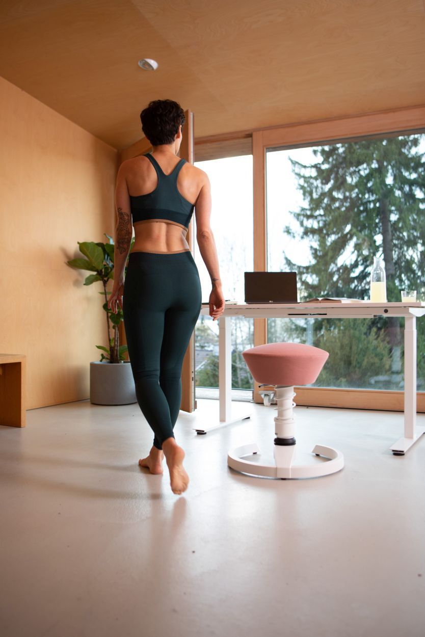 Frau in dunkelgrünem Sport-BH und Leggings, barfuß, vor höhenverstellbarem Schreibtisch und Bürostuhl.