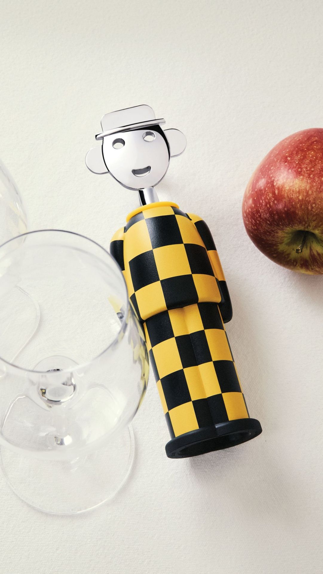 Alessi Alessandro M. Korkenzieher in Gelb-Schwarz mit Schachbrettmuster, neben Weinglas und Apfel.