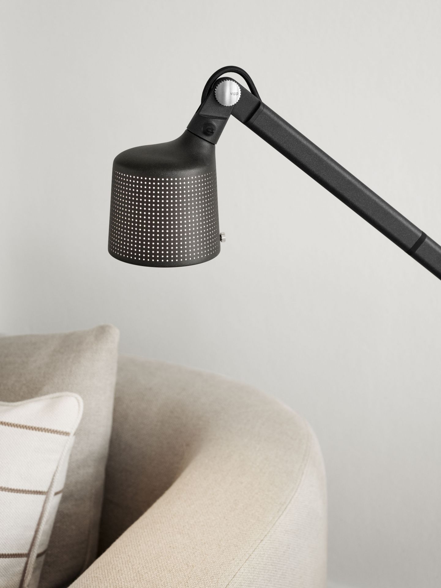 Schwarze Vipp Boden Leselampe mit verstellbarem Arm und perforiertem Lampenschirm neben einem beigen Sessel.