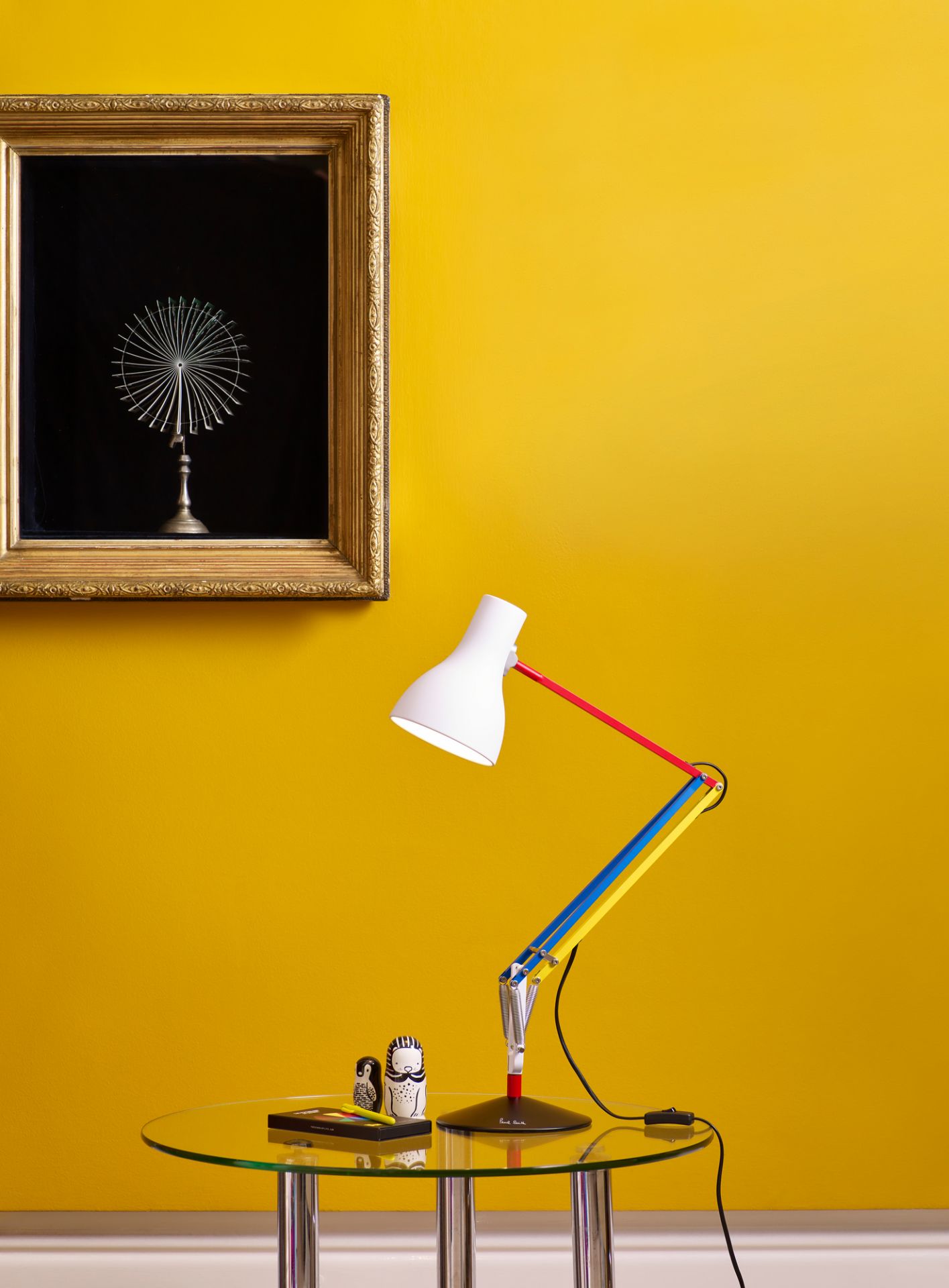 Anglepoise Type 75 Mini Paul Smith Edition Tischleuchte auf Glastisch vor gelber Wand.