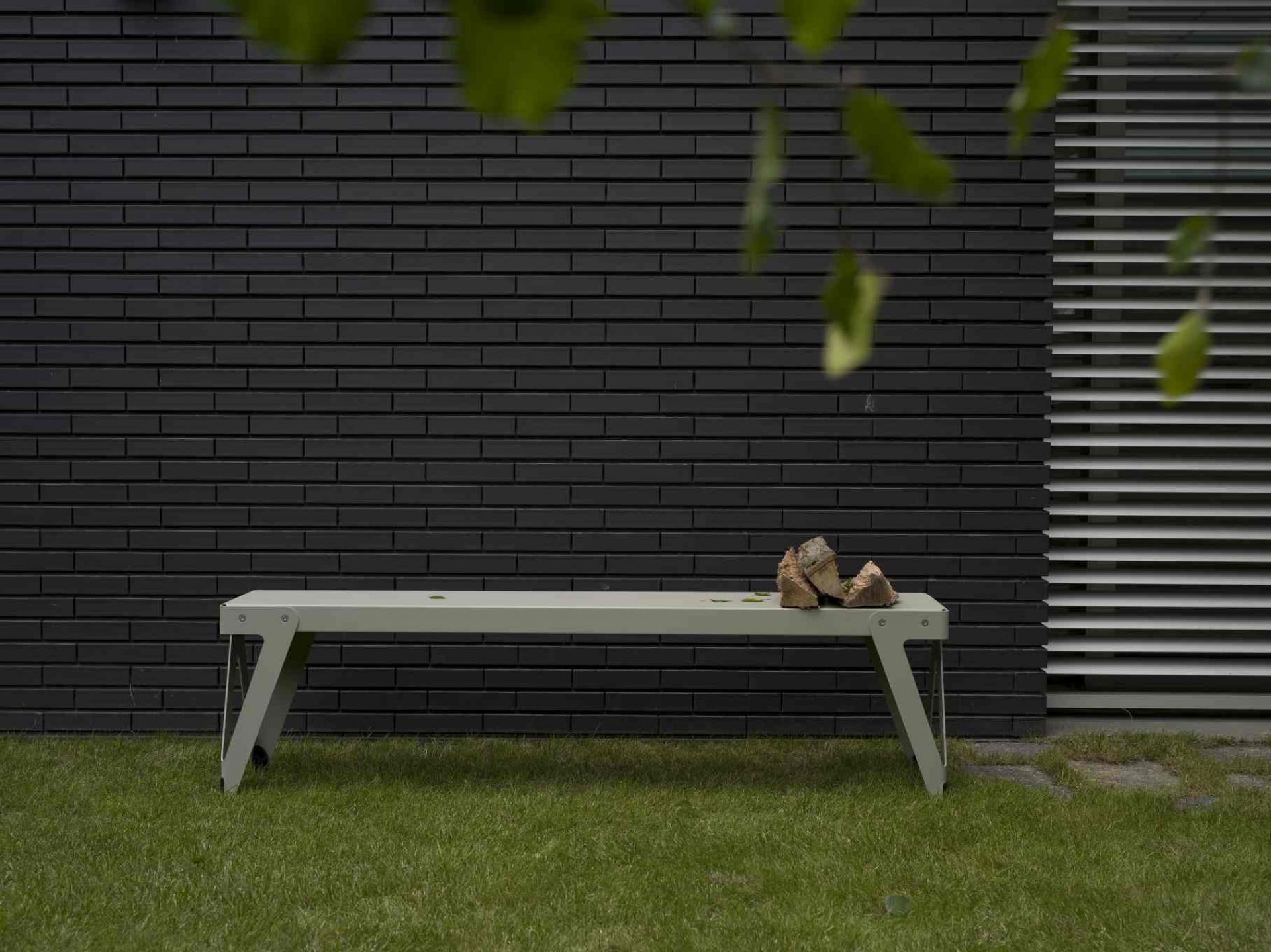 Lloyd Gartenbank: Moderne Outdoor Bank aus Metall vor dunkler Mauer und Rasen.