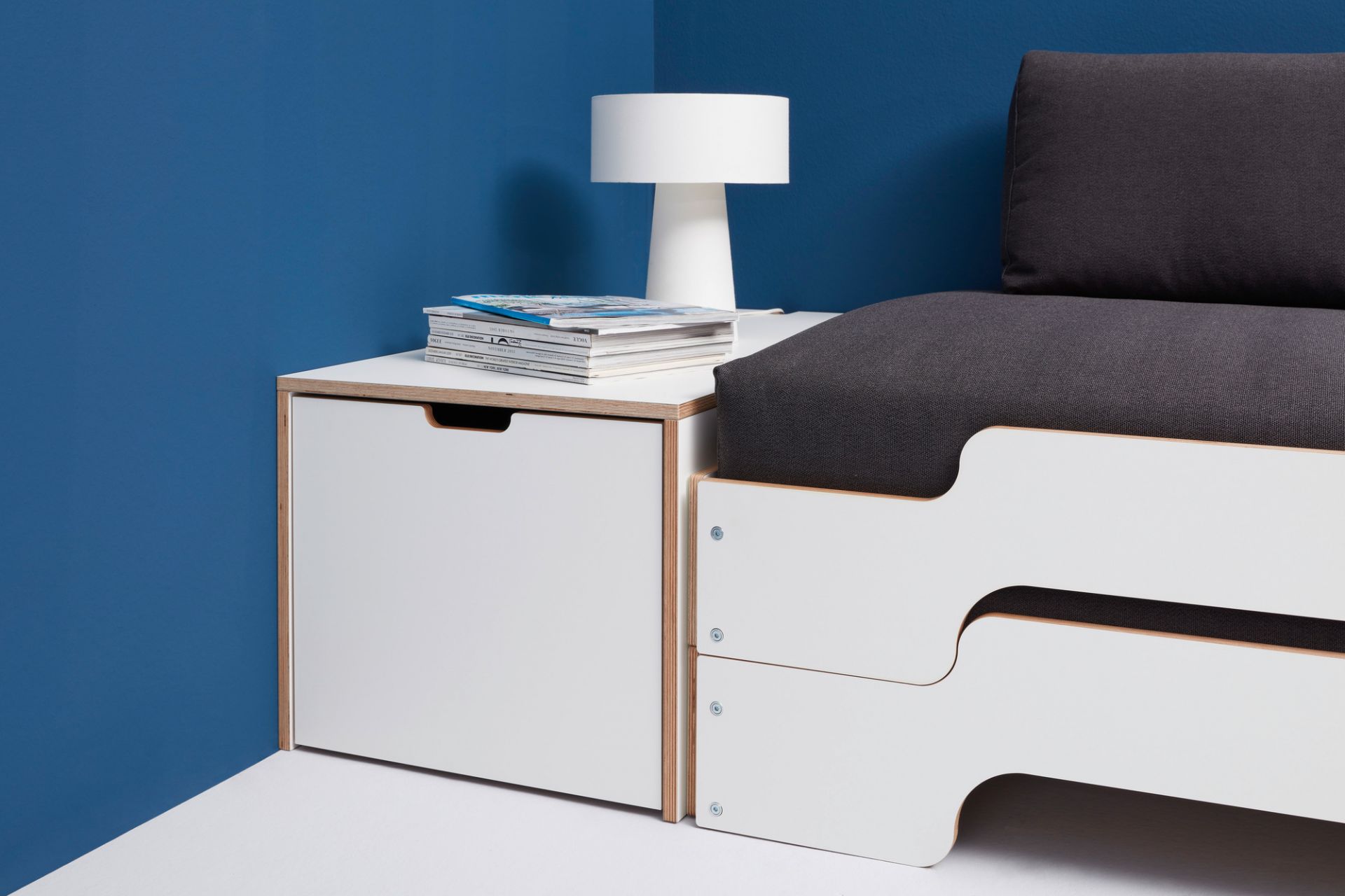 Klassik Bettkasten 16 von Müller Möbelwerkstätten mit Schubkasten, Lampe und Zeitschriften vor blauer Wand.