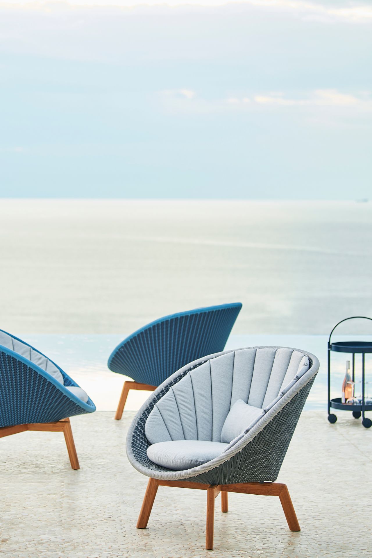 Cane-Line Peacock Loungesessel aus Rattan mit grauen Polstern und Holzbeinen im Außenbereich.
