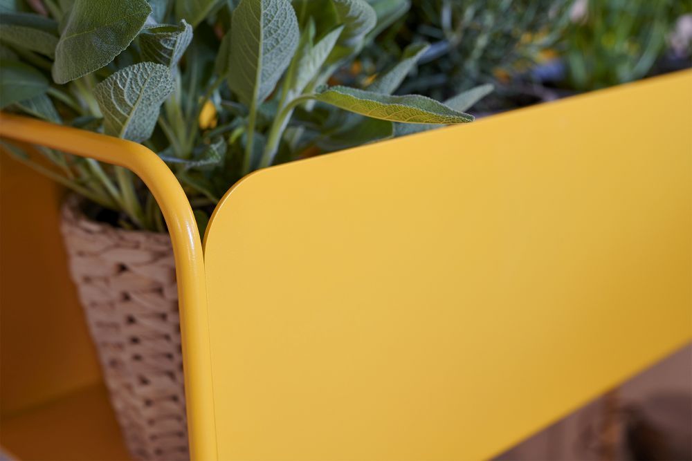 Detailaufnahme: Gelber Fermob Picolino Blumentopf für den Außenbereich mit Pflanze im Korb.
