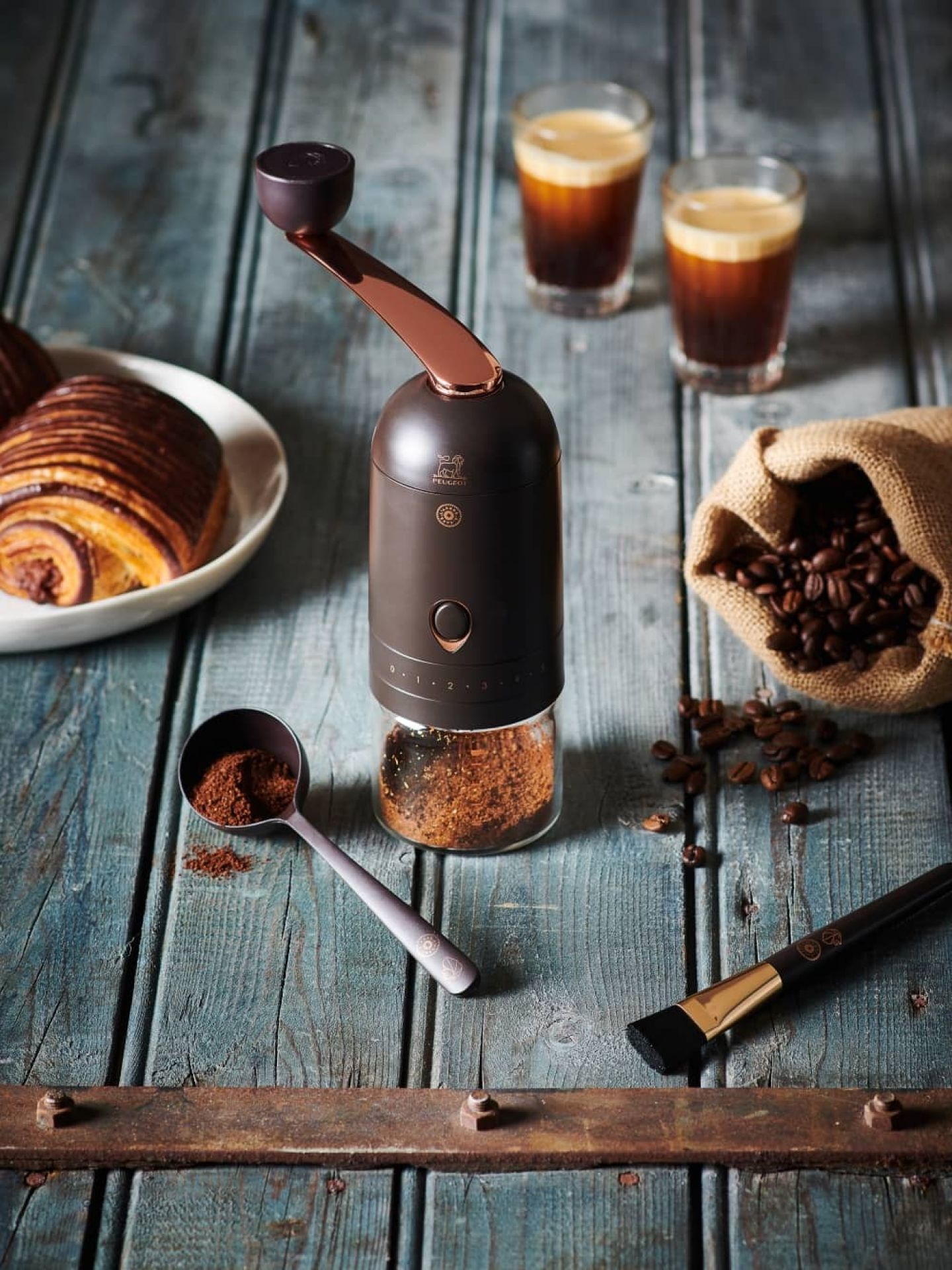 L'Arbre à Café Peugeot Kaffeemühle mit frisch gemahlenem Kaffee, Espresso und Gebäck auf rustikalem Holztisch.
